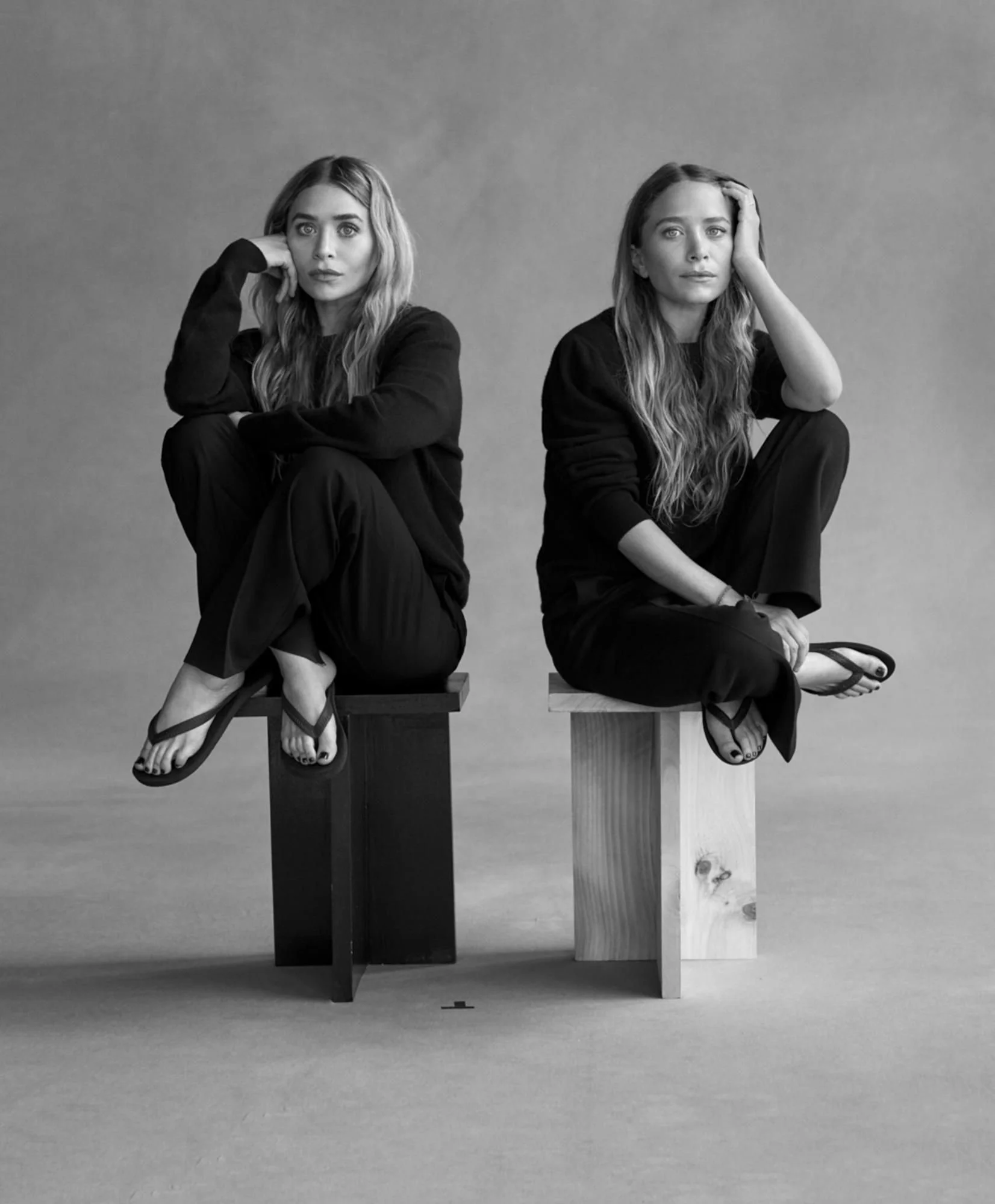 Black-and-white editorial photo featuring two women seated on minimalist wooden stools, wearing relaxed monochrome outfits and sandals. Clean, quiet-luxury styling representing the understated, elevated aesthetic of luxury label The Row.