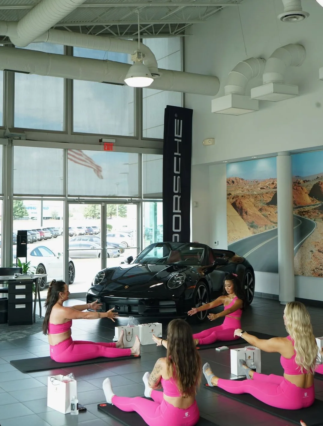 Pilates class hosted inside a Porsche dealership, women in pink matching activewear stretching in front of a black Porsche sports car during luxury fitness brand collaboration.