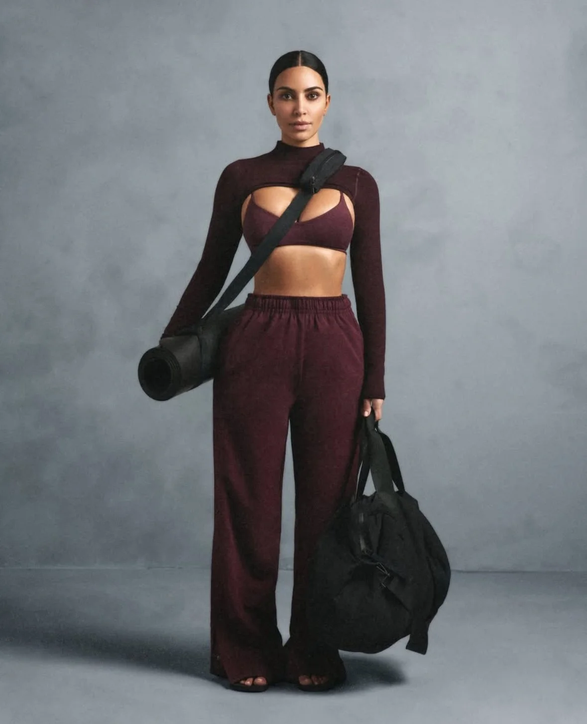 Kim Kardashian wearing burgundy Skims activewear set with cut-out top, holding yoga mat and gym duffel bag, shot against neutral studio backdrop.