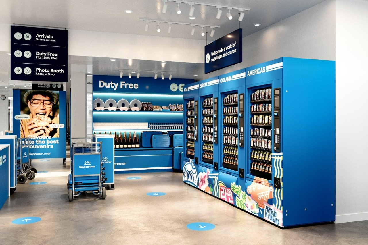 Immersive “airport-style” retail pop-up with snack vending machines labeled by global regions, luggage carts, and directional signage. Branded in blue with playful travel-themed messaging.
