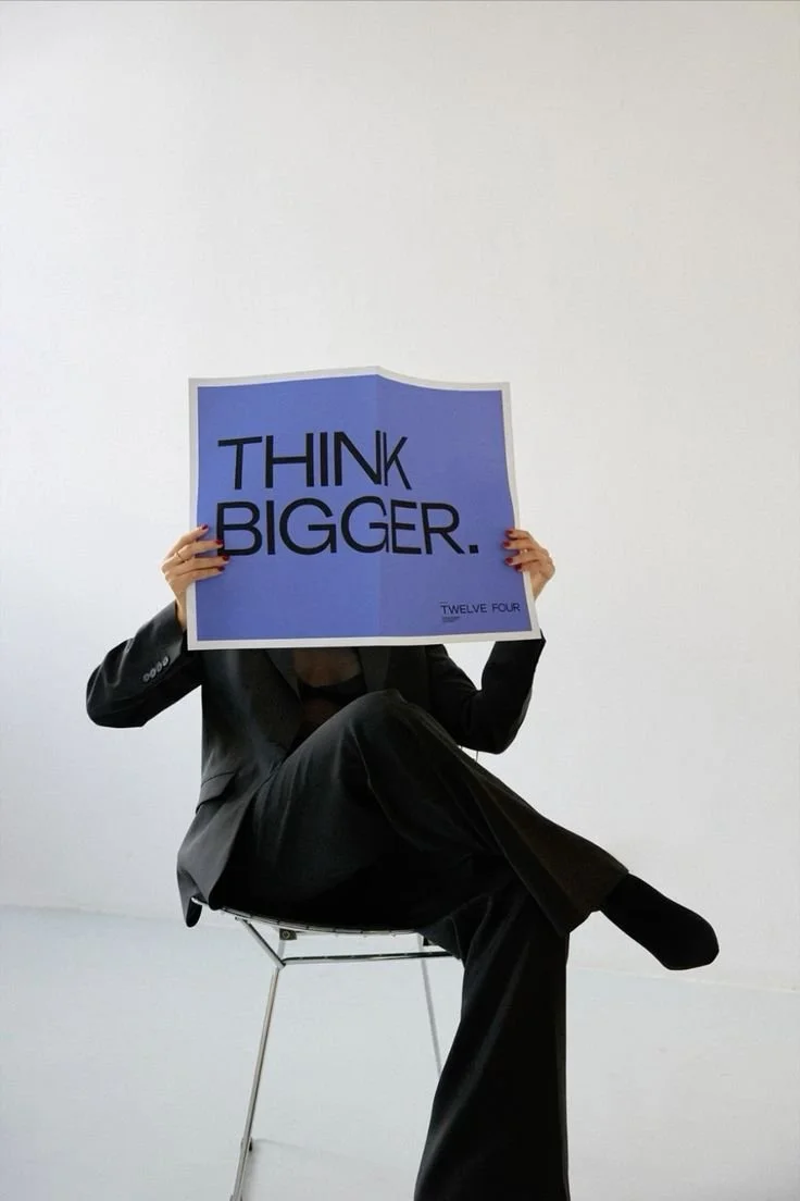 A person holding a newspaper stating "think bigger" siting crossed legged on a chair.