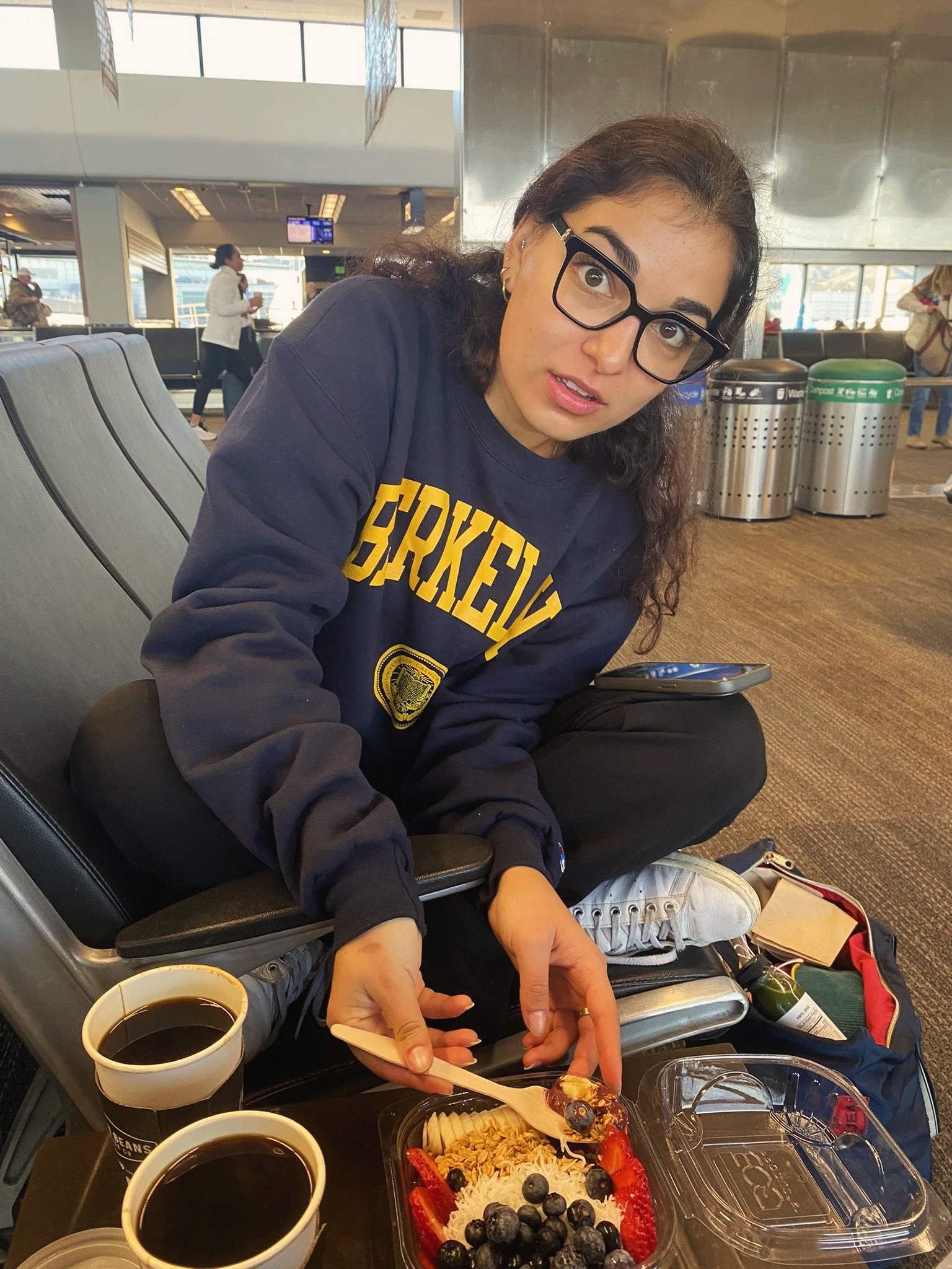A woman with glasses sitting in an airport holding a fork with food, with two cups of coffee, sitting next to a container of fruit and granola, in an airport terminal.