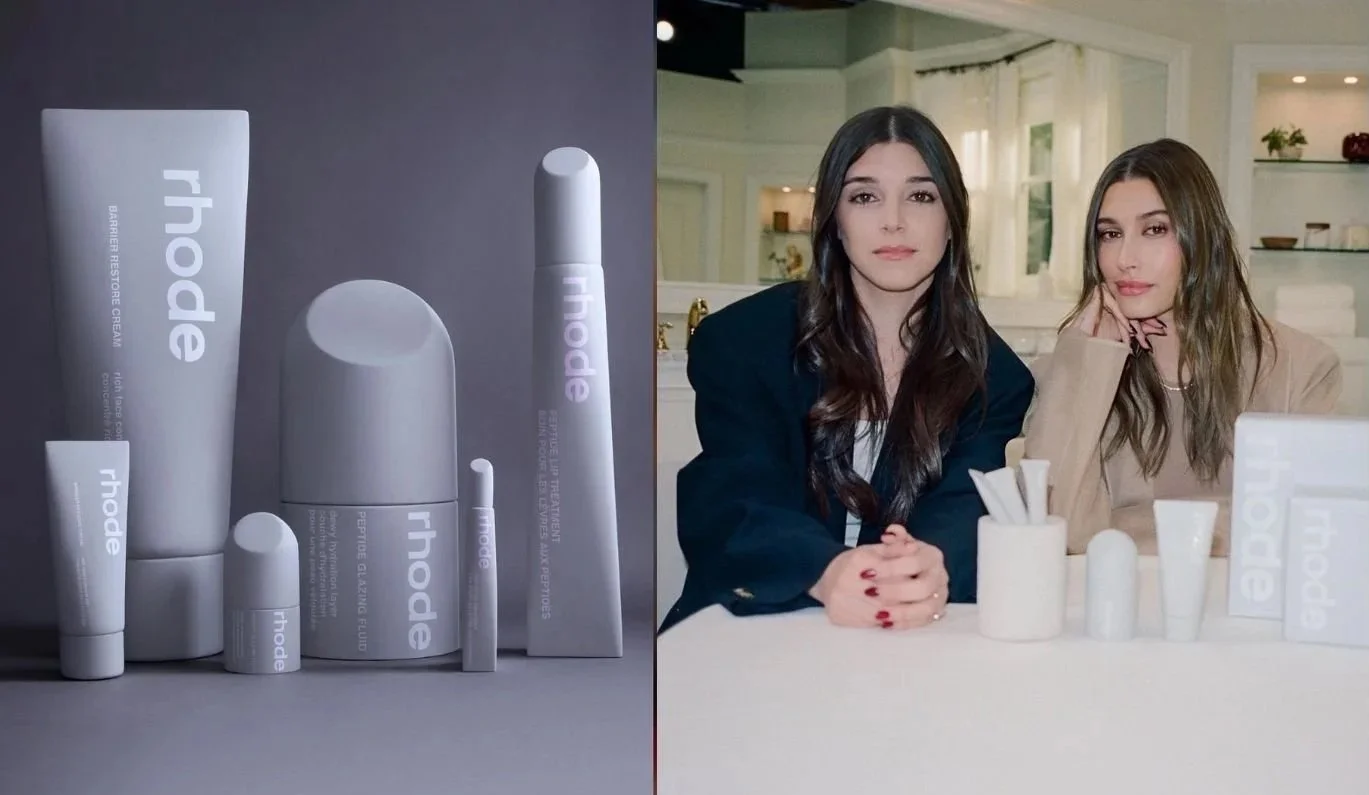 Rhode skincare founders sit beside the brand’s minimalist grey packaging lineup, featuring the Peptide Glazing Fluid, Barrier Restore Cream, and Lip Treatment — symbolizing Rhode’s clean design and strategic brand identity