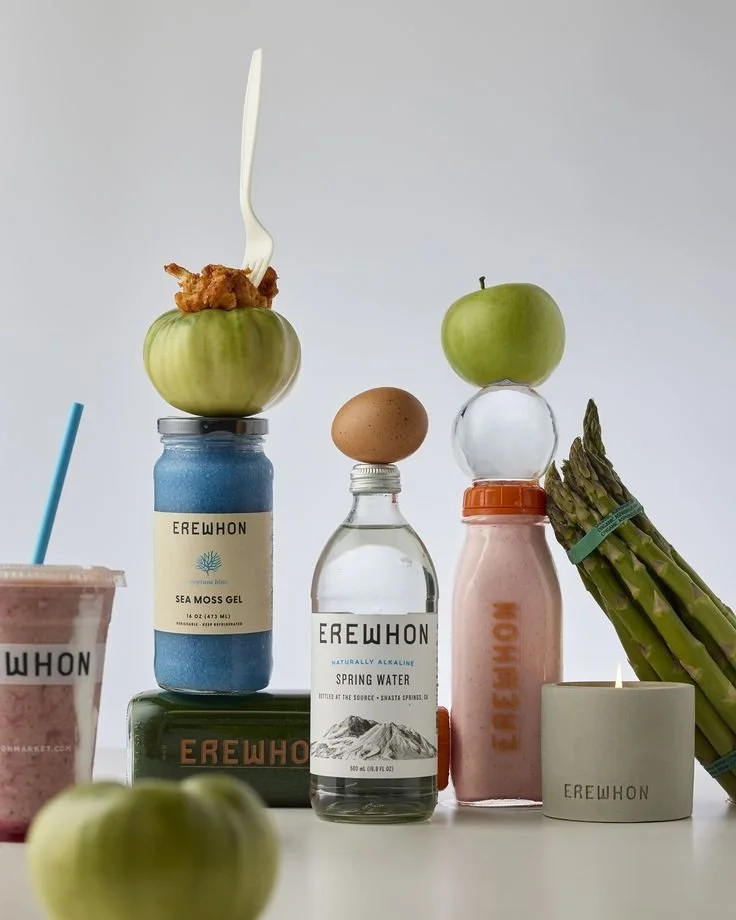 An artistic, minimalist studio shot featuring a vertical stack of Erewhon-branded products and fresh produce against a clean white background.