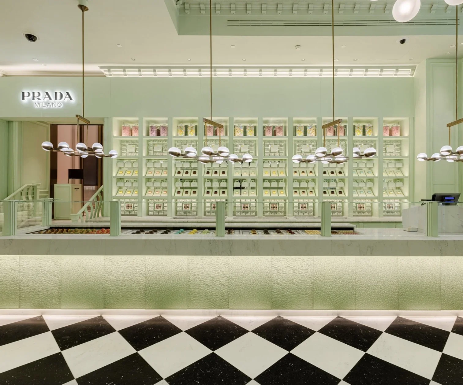 An interior view of a mint-green Prada Milano boutique and pastry shop, featuring illuminated glass display shelves, a long white marble counter, modern hanging globe light fixtures, and a black-and-white checkered floor.