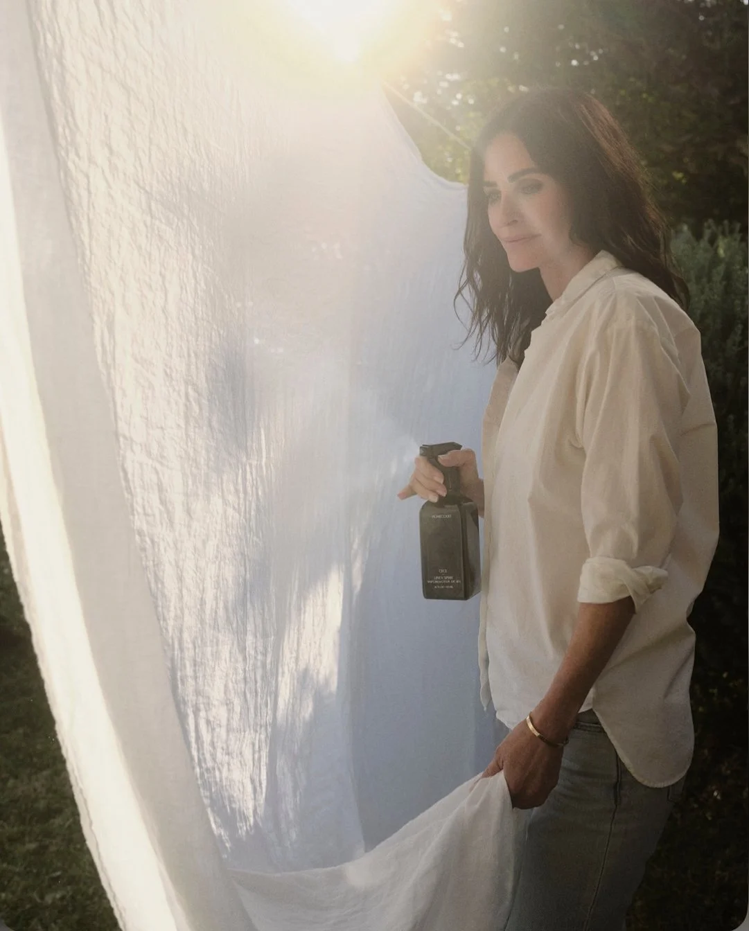 Courteney Cox gently misting a linen spray on white sheets outdoors, illuminated by soft natural sunlight