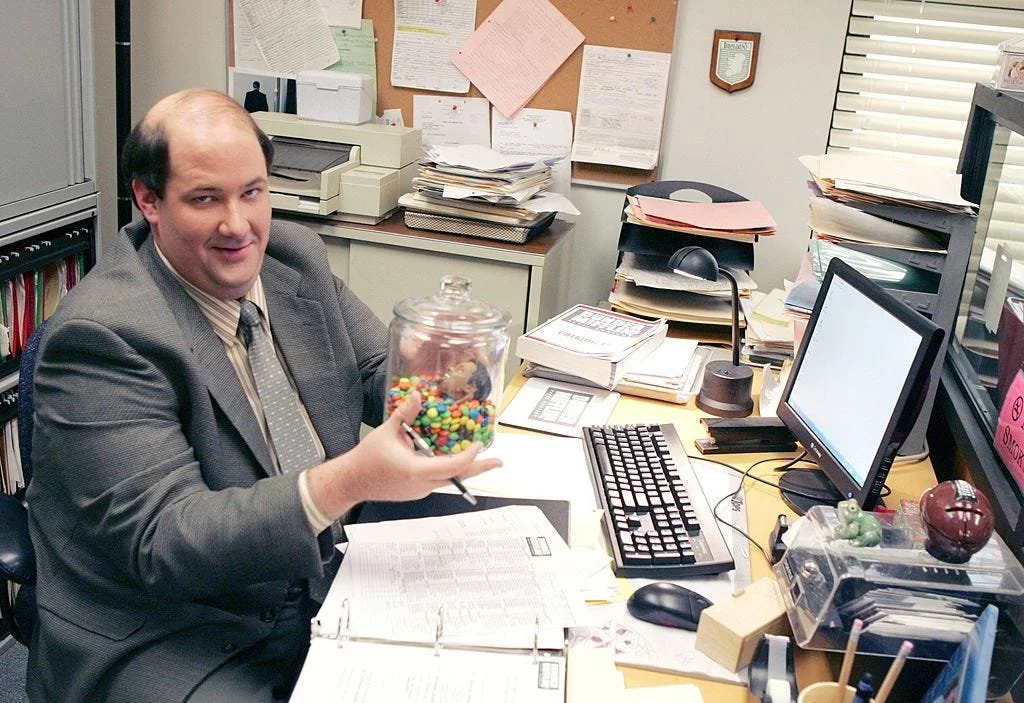 Brian Baumgartner as Kevin Malone sitting at his cluttered office desk in The Office, holding a jar of M&M’s, portraying his iconic accountant character known for humorous financial mistakes.