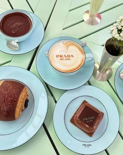 A top-down view of a light green table set with pastel blue Prada Milano tableware, featuring a latte with "PRADA MILANO" foam art, a chocolate-glazed pastry with a Prada plaque, a pain au chocolat, hot chocolate, and a small vase of white flowers.
