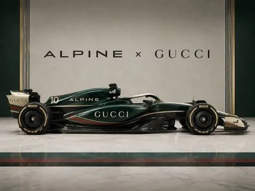 A side-profile view of an Alpine x Gucci Formula 1-style racing car in dark green with cream and gold branding, featuring prominent "ALPINE" and "GUCCI" lettering against a minimalist wall with the text "ALPINE x GUCCI".