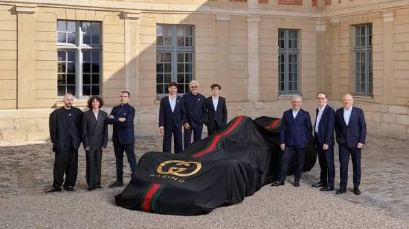 A group of seven people in dark suits posing in a historic stone courtyard next to a racing car covered by a black satin sheet featuring red-and-green stripes and a large gold Gucci Racing logo.