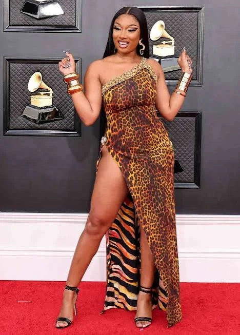 Megan Thee Stallion posing on a red carpet wearing a one-shoulder animal-print gown with a high leg slit, gold statement cuffs, and black strappy heels, with Grammy award wall plaques in the background.