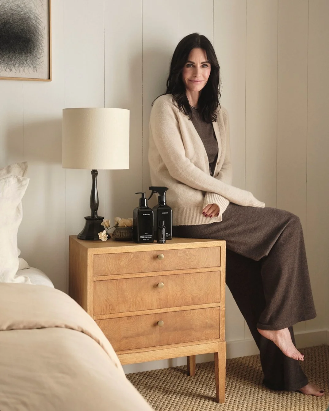 Courteney Cox seated on a bedside table in a warm, minimalist bedroom with Homecourt products displayed beside her.