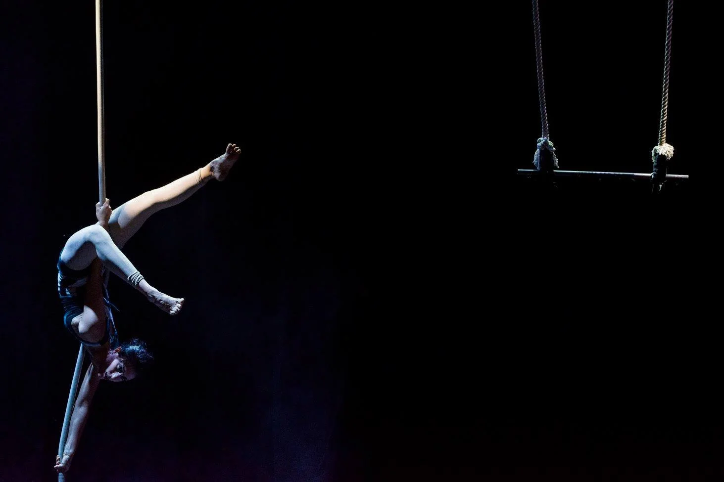 Aerialist on rope in a one-arm split, empty trapeze hanging nearby in the dark