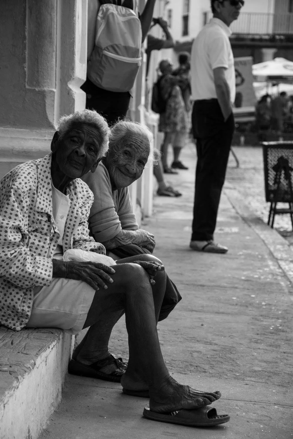 שתי חברות מבוגרות יושבות על מדרכה בהוואנה ומחייכות למצלמה