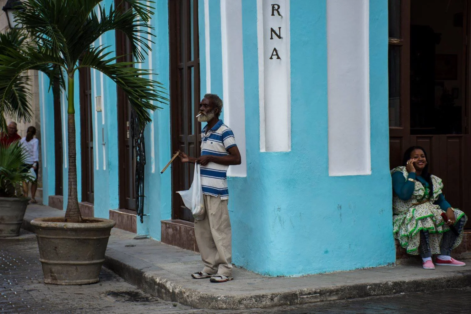 איש מבוגר נשען על מקל מחוץ לבניין טורקיז בעוד אישה בשמלה צבעונית יושבת בפתח הדלת
