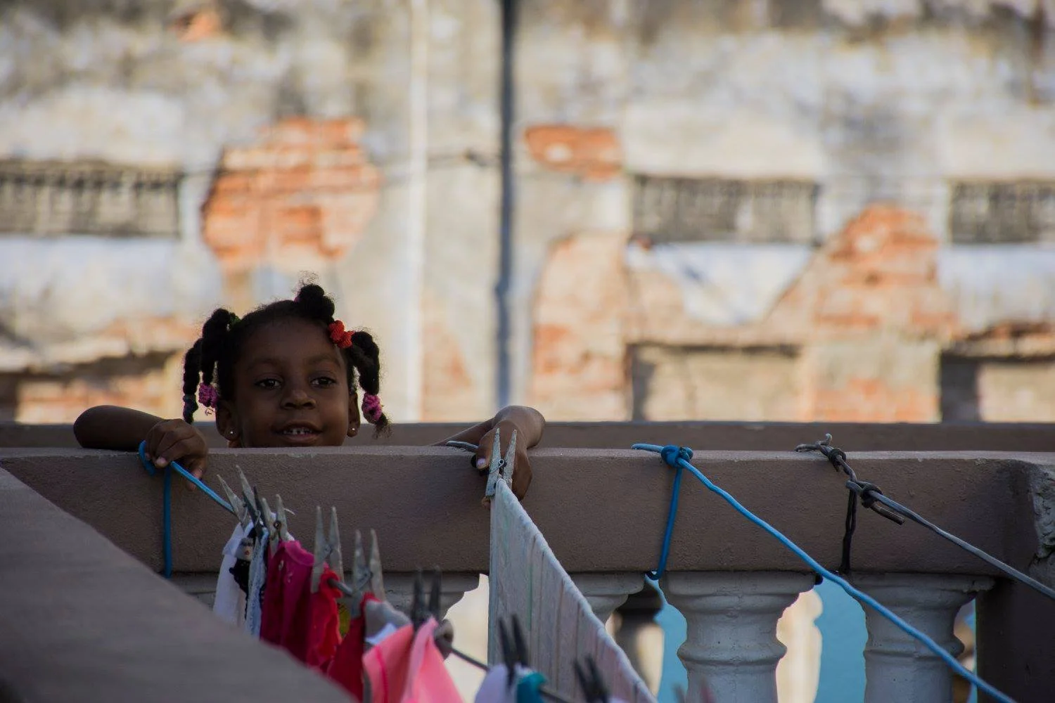 ילדה קטנה עם צמות מציצה מעל מעקה מרפסת כשכביסה צבעונית תלויה על חבל