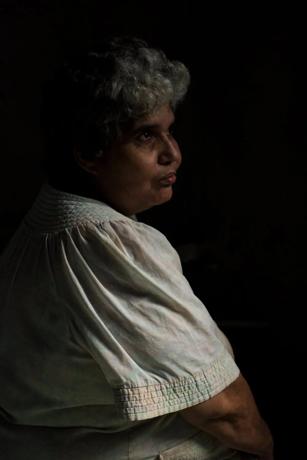 אישה בפרופיל, אור חלון רך על פניה בחדר חשוך, חולצה לבנה ומבט מהורהר