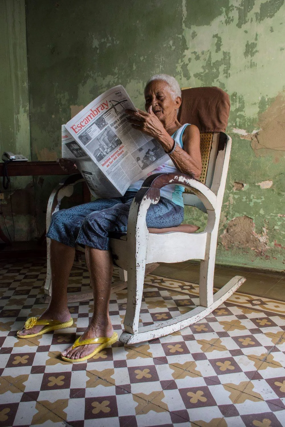 אישה מבוגרת בכיסא נדנדה לבן קוראת את העיתון Escambray בחדר ירקרק דהוי עם רצפת אריחים ונעלי אצבע צהובות