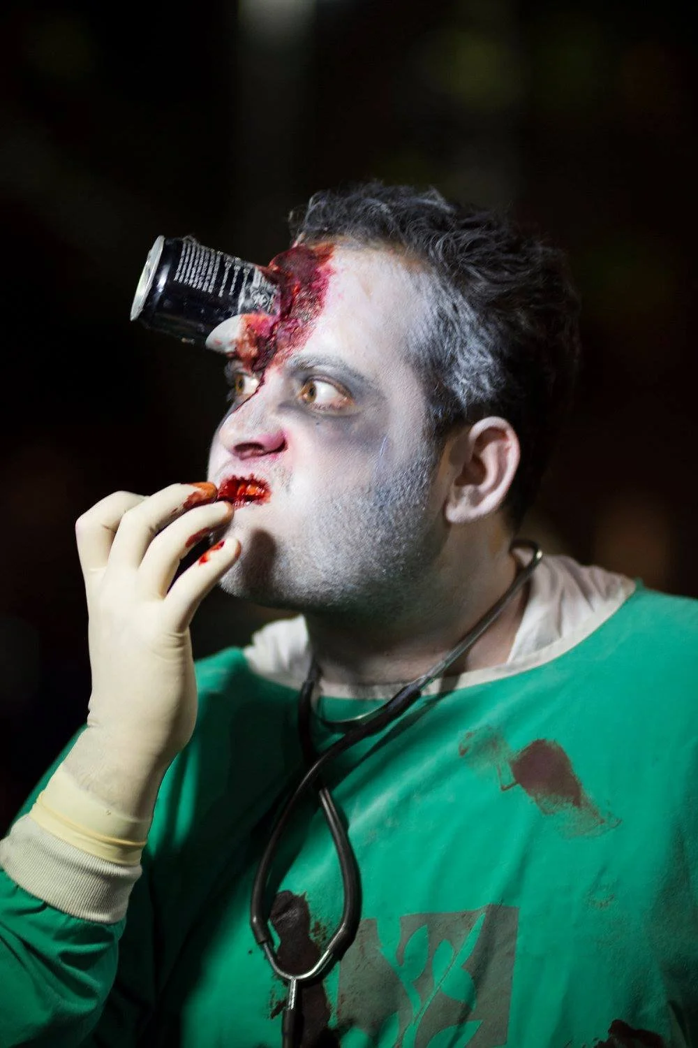 גבר במדי רופא ירוקים קרועים עם איפור מדמם ועדשת מצלמה מזויפת תקועה במצח, מצולם באמצע ביס