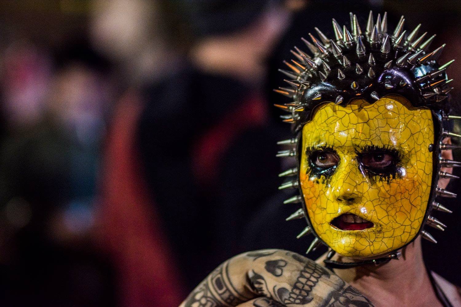 איש עם מסכה צהובה סדוקה ויתדות מתכת, הכתפיים המקועקעות חשופות