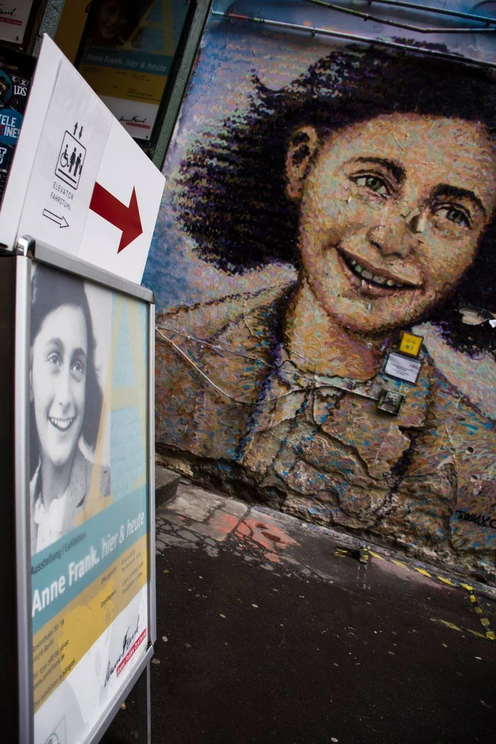 ציור קיר של אנה פרנק עם שלט מוזיאון וחץ אדום בכניסה, מדרכה רטובה