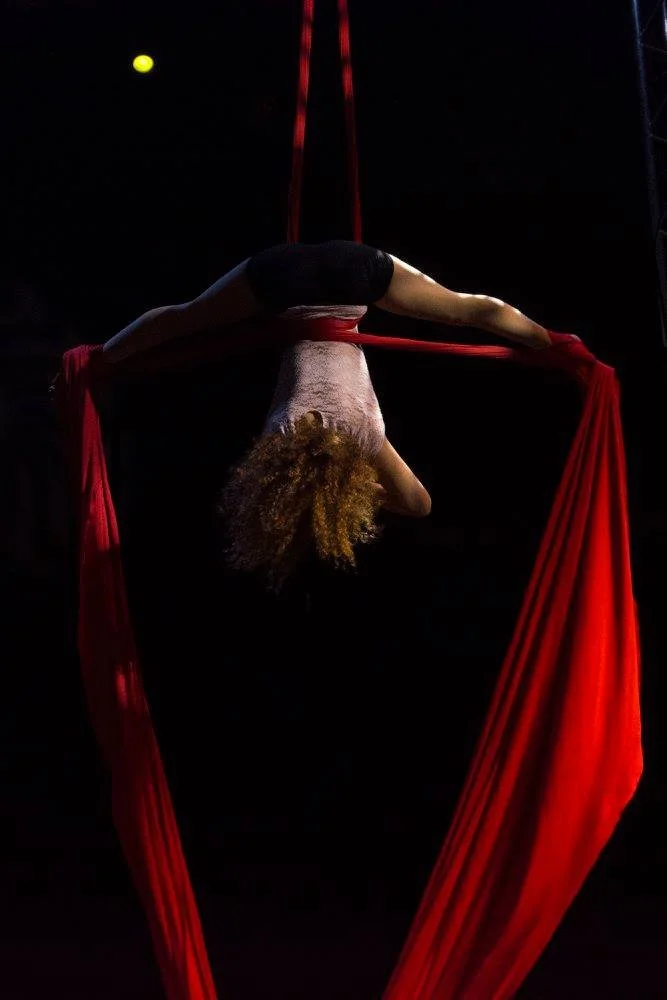 אקרובטית אוויר מתקשתת לאחור מעל בדים אדומים בוהקים, השיער תלוי בתוך האור