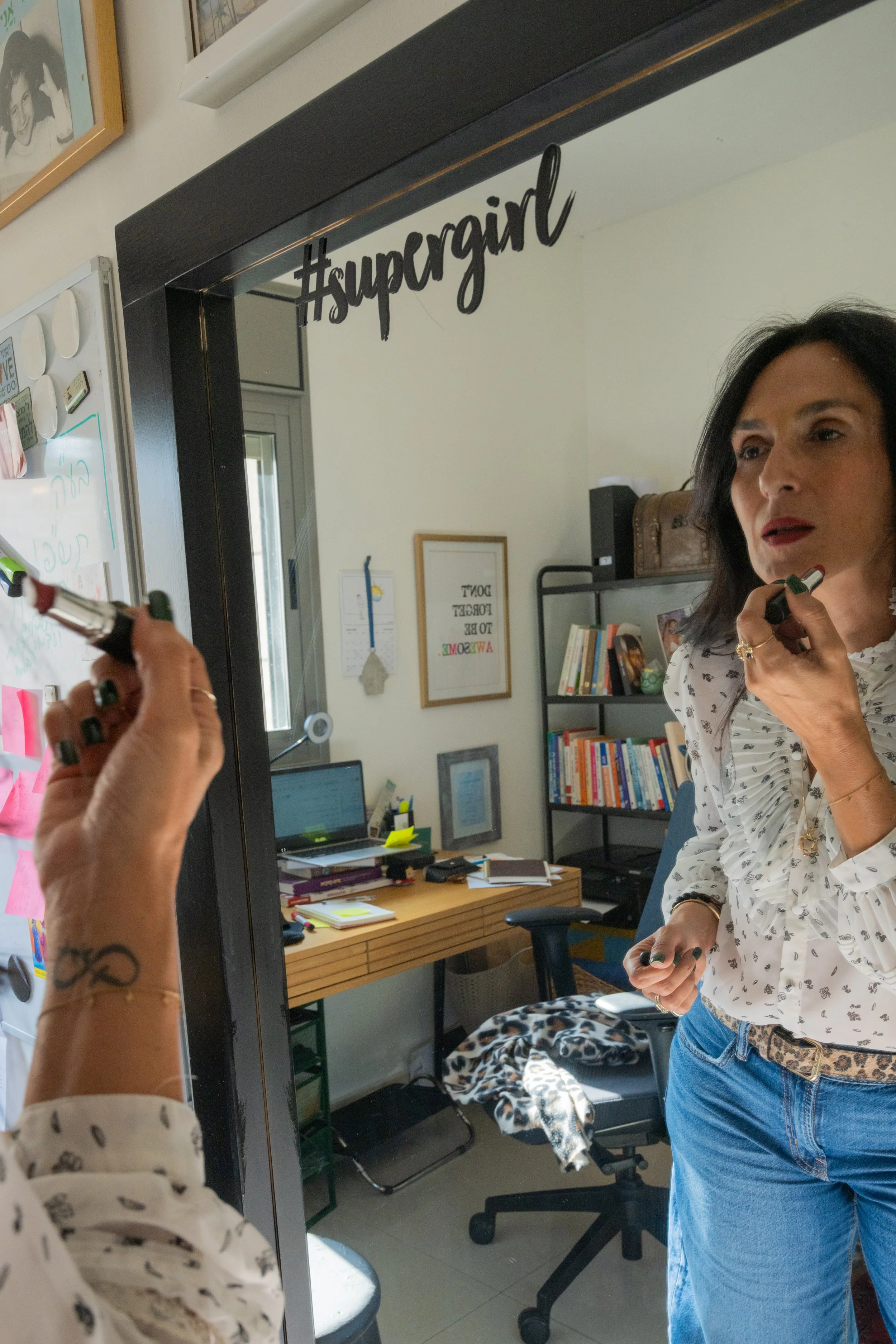 Limor Poran puts on red lipstick in front of a mirror with the word “#supergirl” above it, getting ready for another workday in her home office.