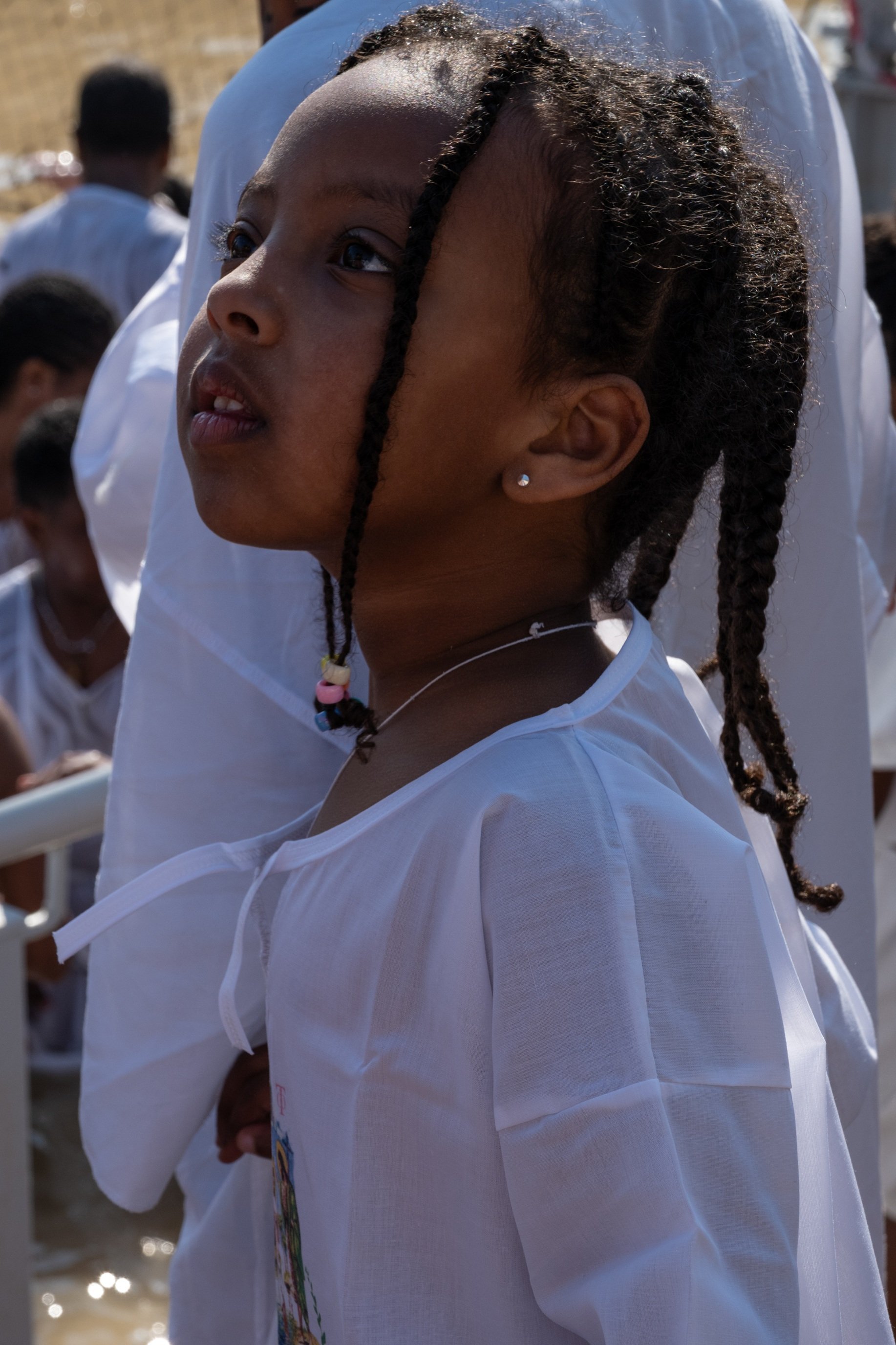 Close-up of a girl in a white robe looking upward, sunlight on her face, people in white behind her