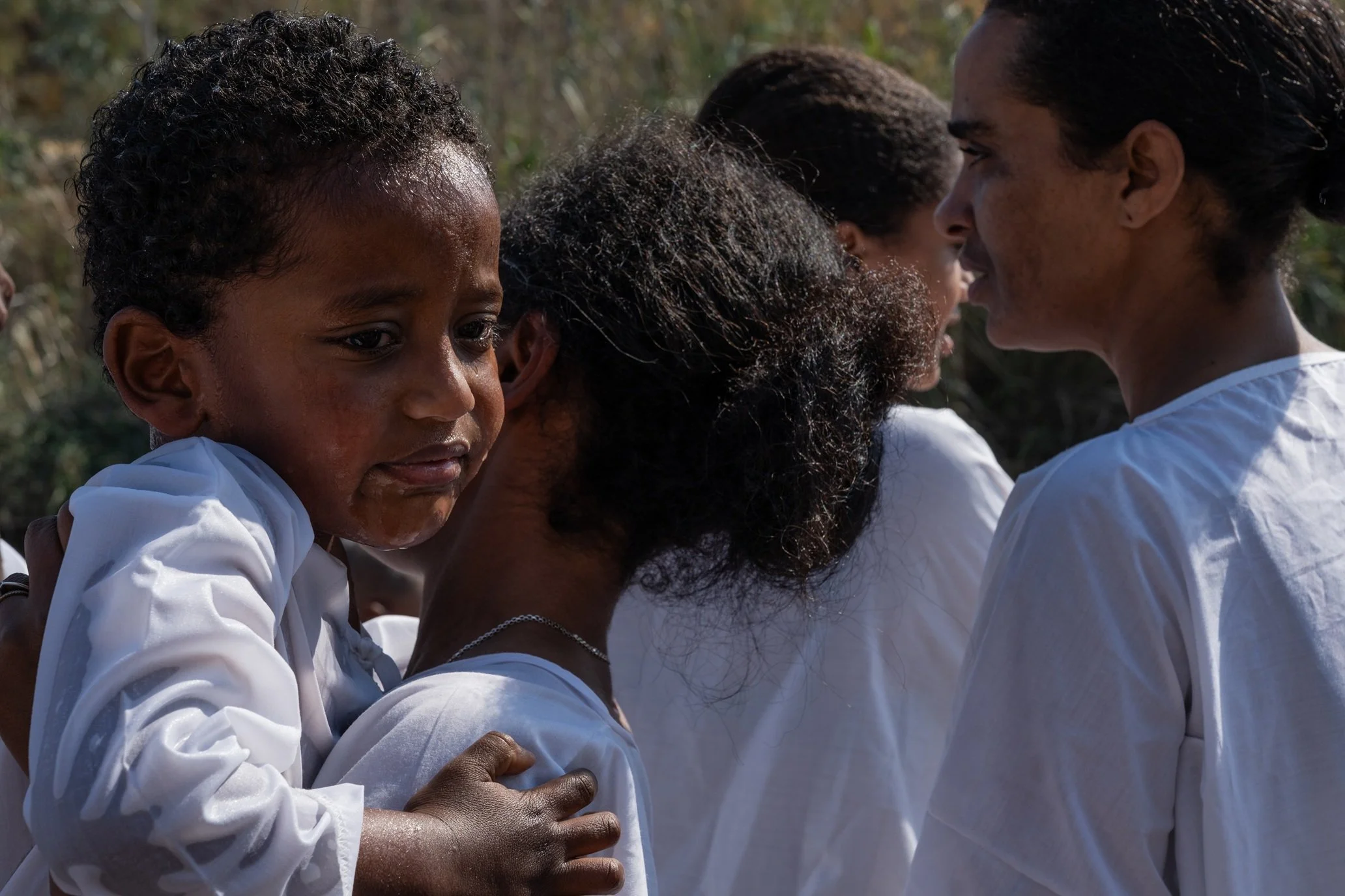 Crying child in a wet white robe being held close by an adult in the crowd near the river