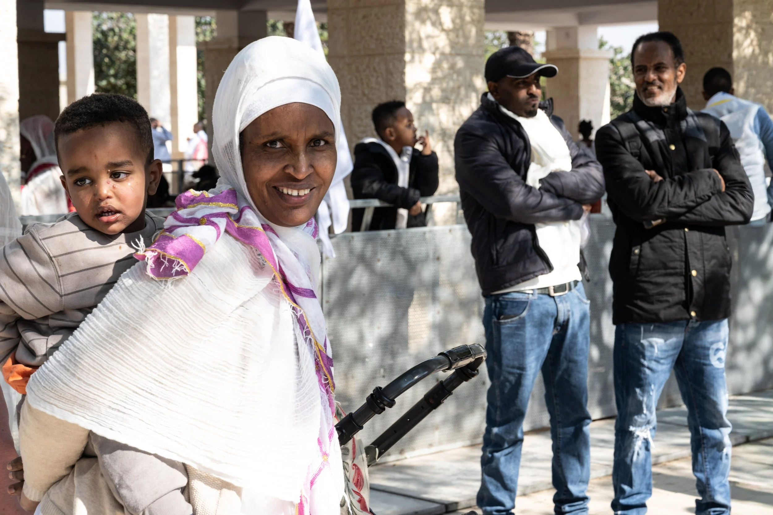 Smiling woman in a white shawl carrying a toddler on her back, with onlookers in the background