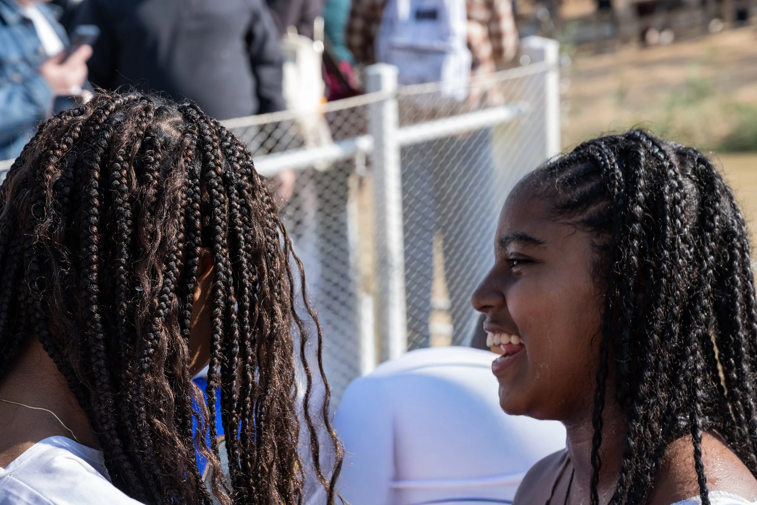 Two girls in profile, smiling as they look toward the river, with bright light and the crowd around them
