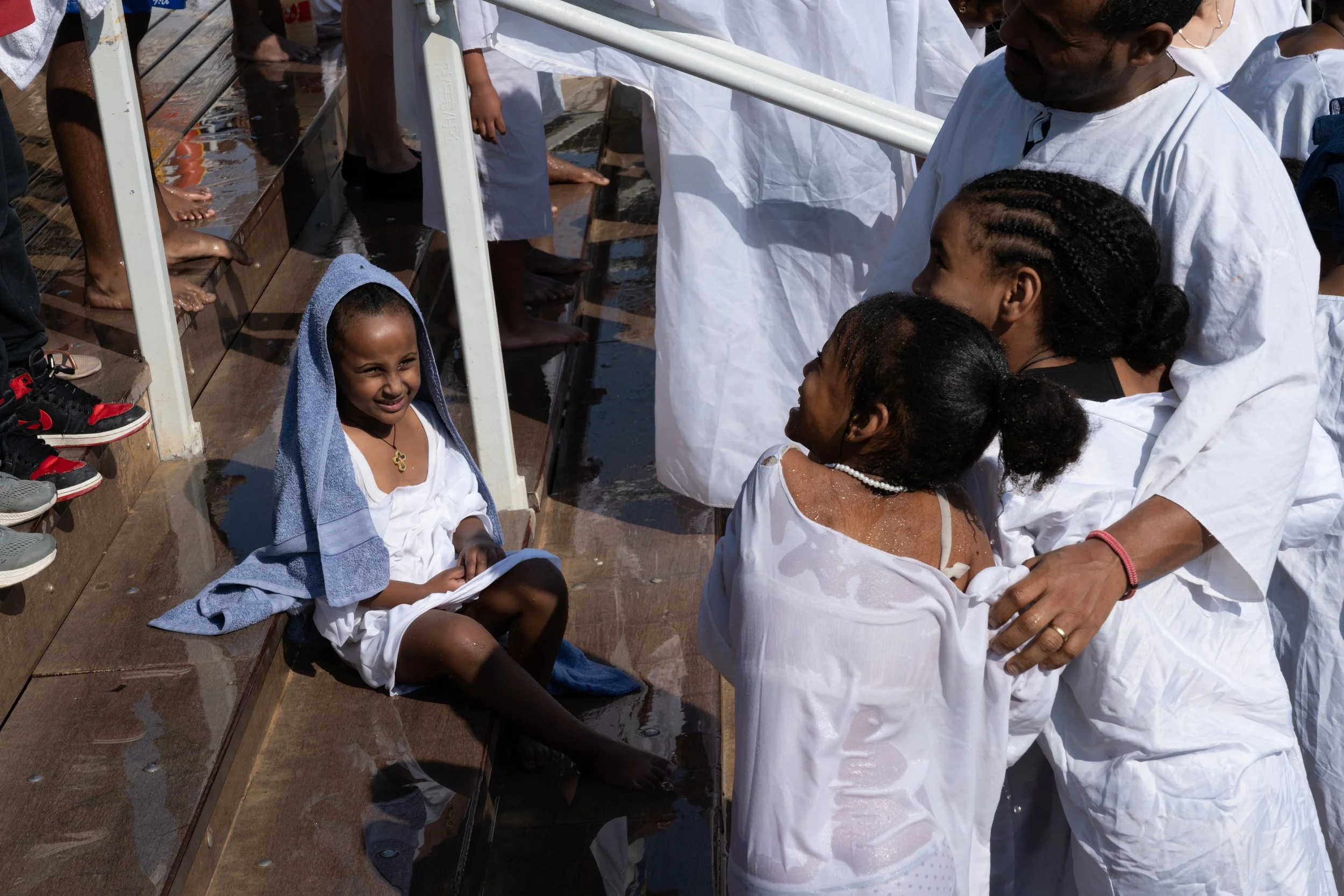 Children in wet white clothes gathered on the wooden deck by the river, two children held close by an adult