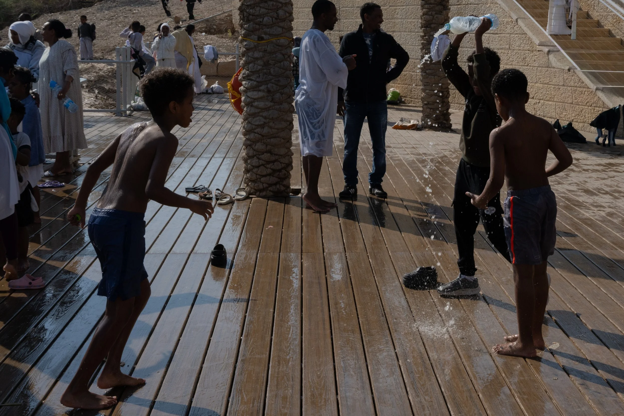 Children playing barefoot on the wooden deck near the water, with people watching from behind