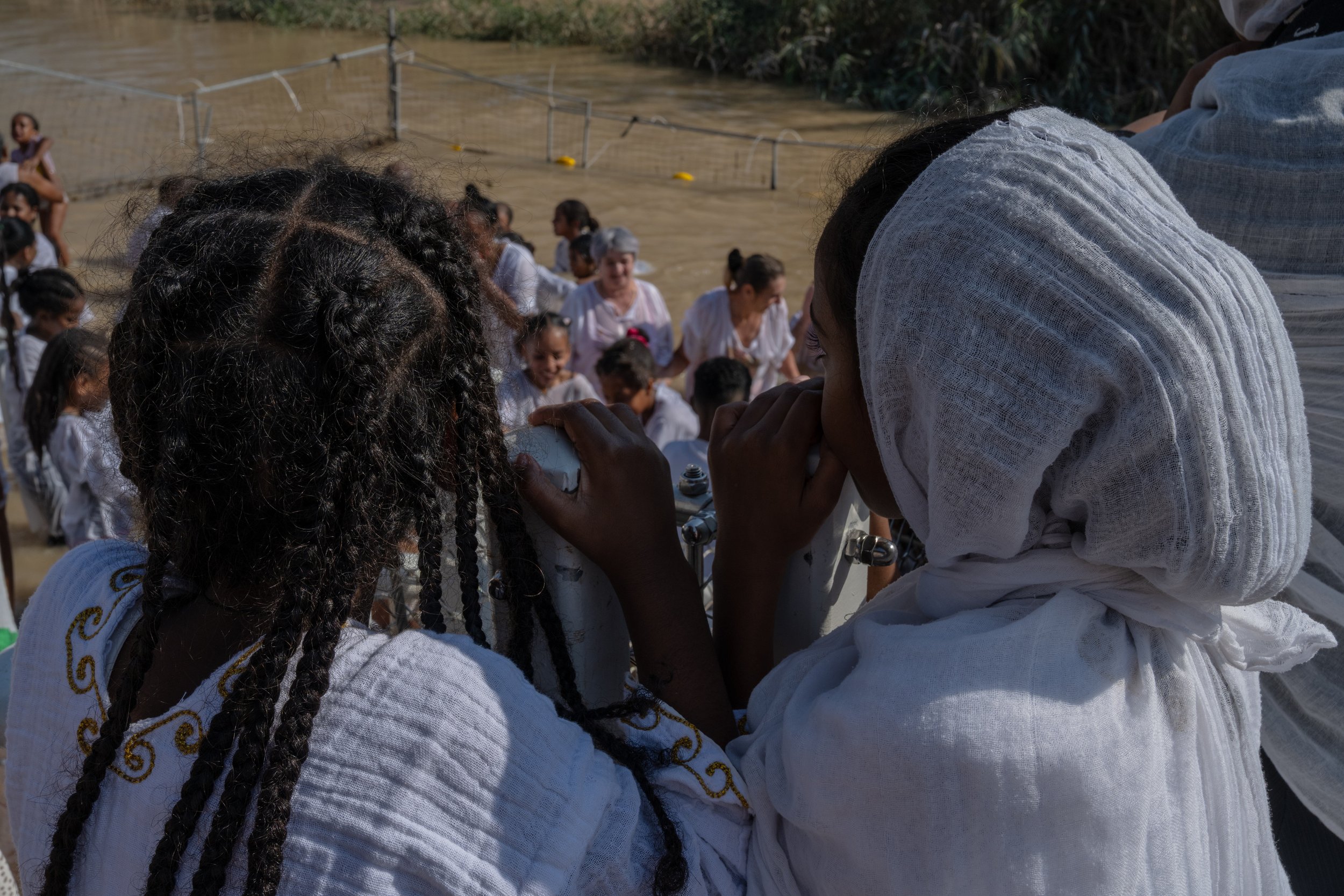 Two girls from behind, heads close together, wearing white shawls as they look toward the river