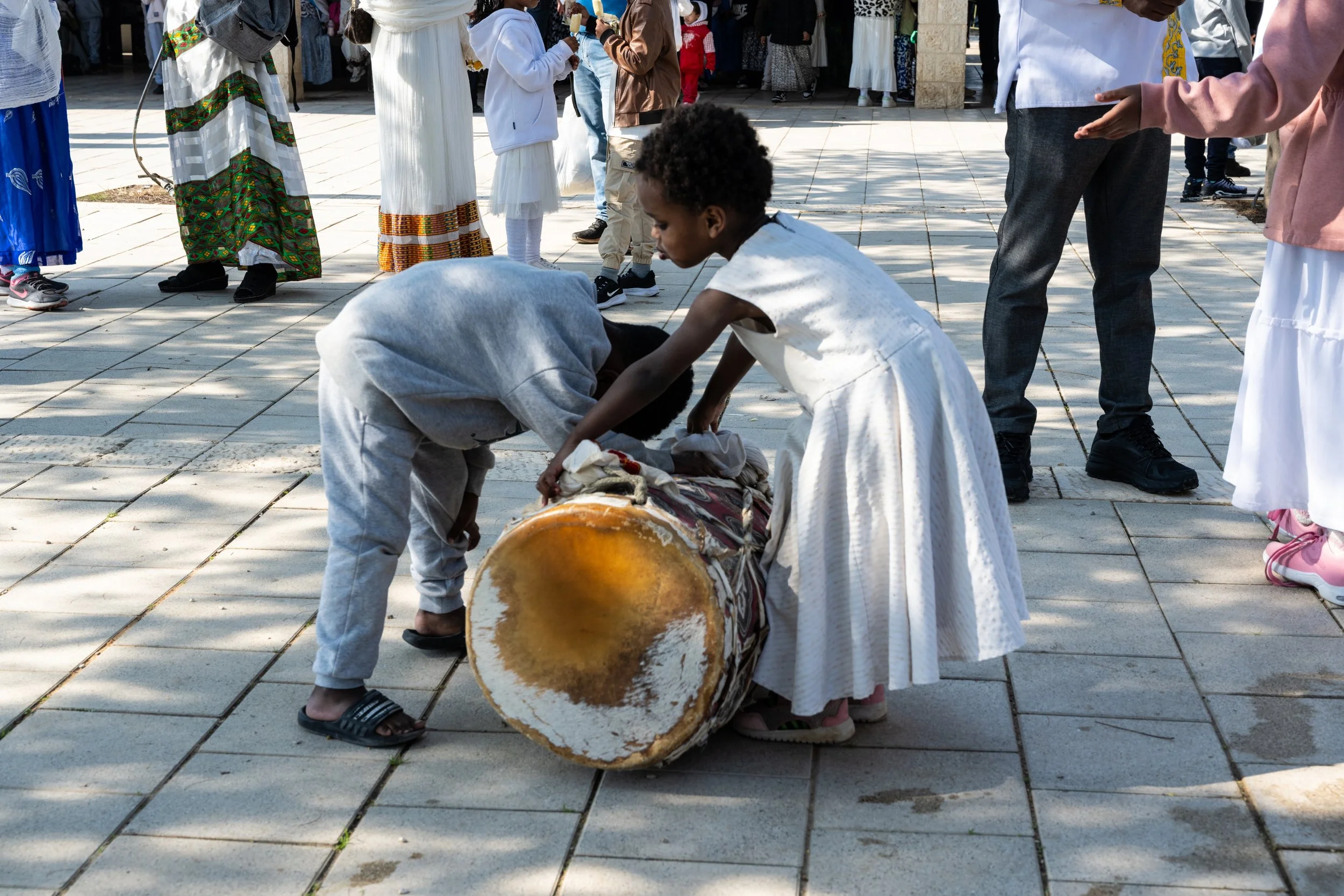 Children lifting and steadying a large drum on the plaza, surrounded by families in white