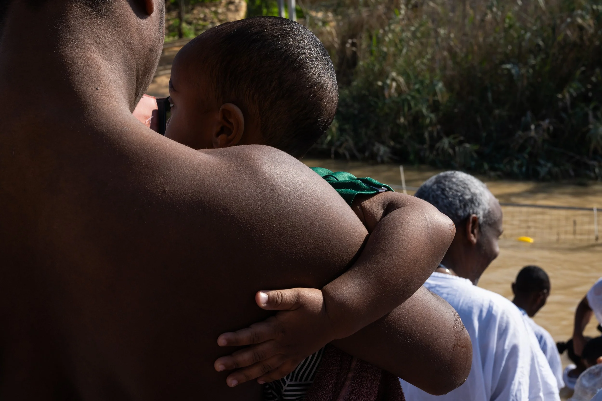 Small child hugging an adult’s shoulder, seen from behind, with the muddy river and pilgrims in white