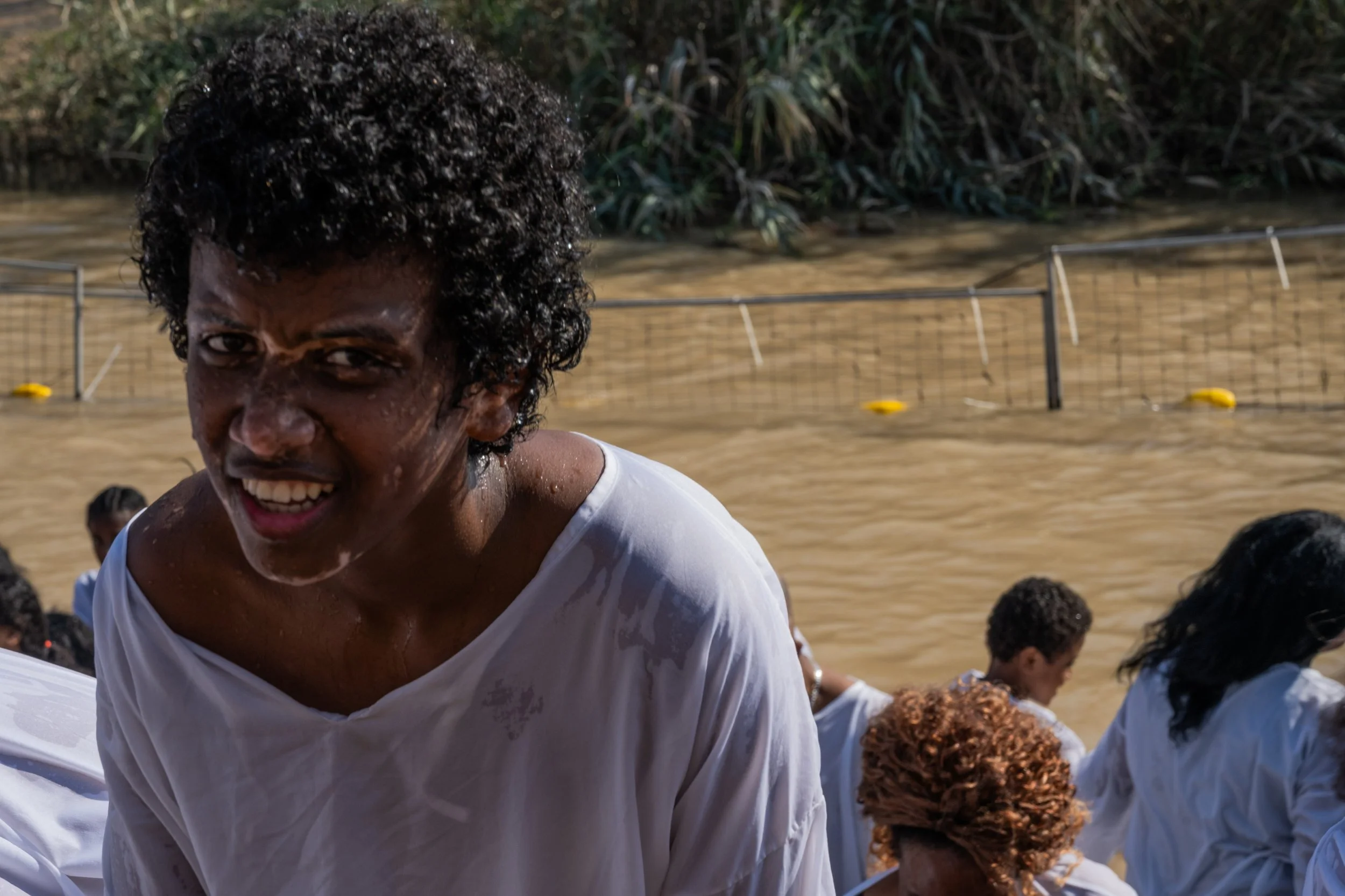 Wet young man in a white shirt smiling near the Jordan River, with the river and crowd behind him