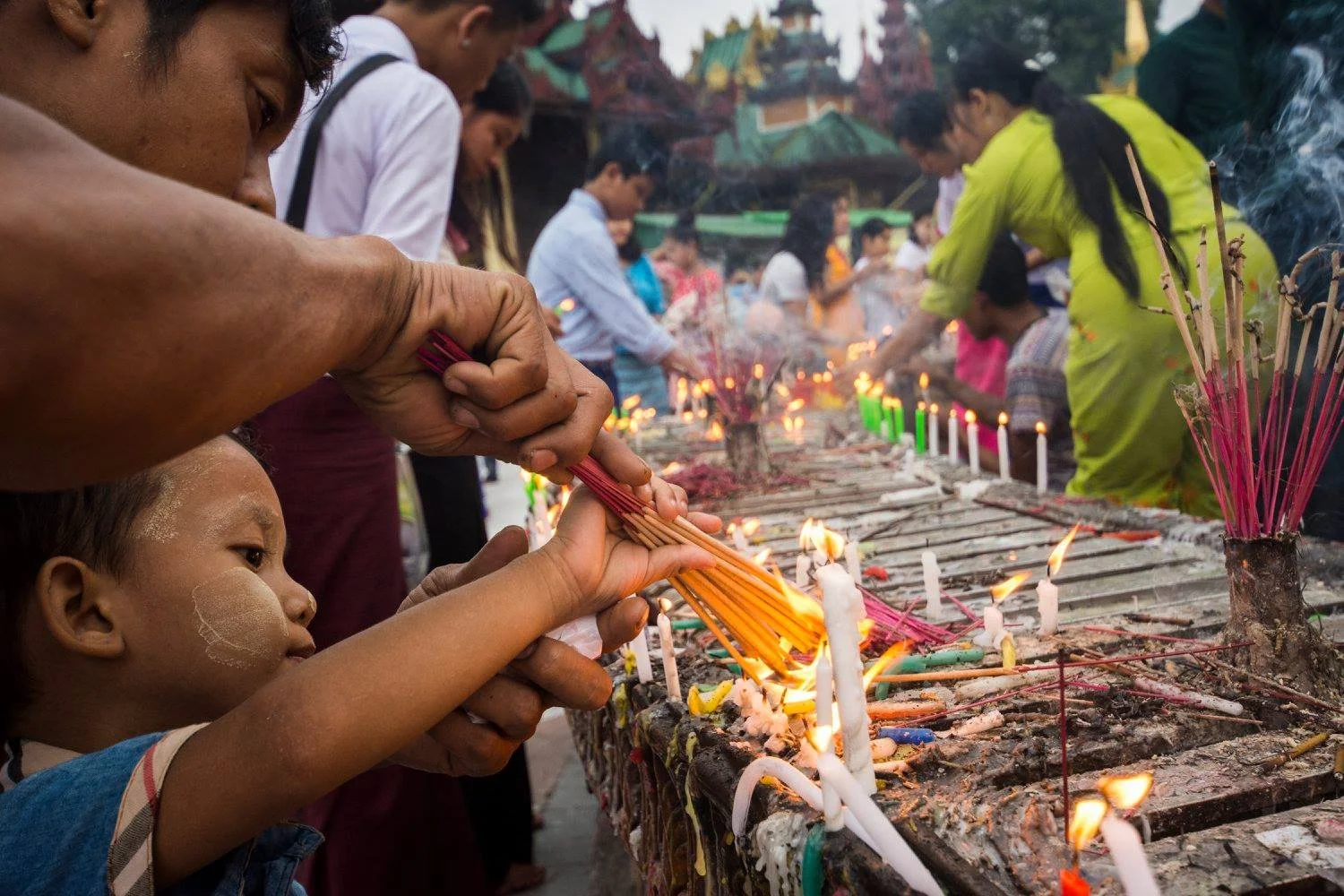 Adult hands helping a young child place incense and candles among many small flames during the Thadingyut Festival in Myanmar