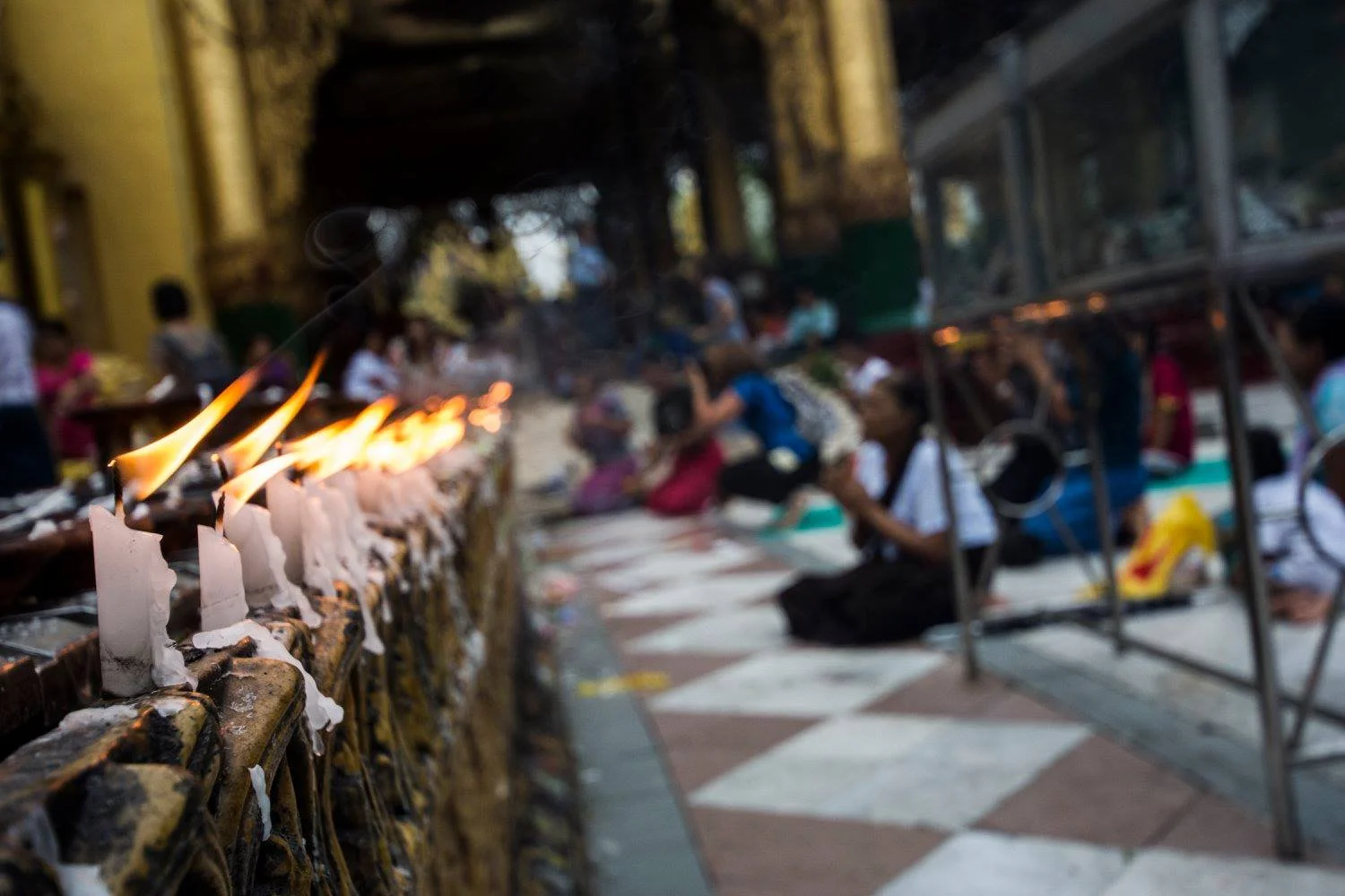 Row of burning candles in sharp focus with worshippers sitting and praying in the background at a pagoda in Myanmar