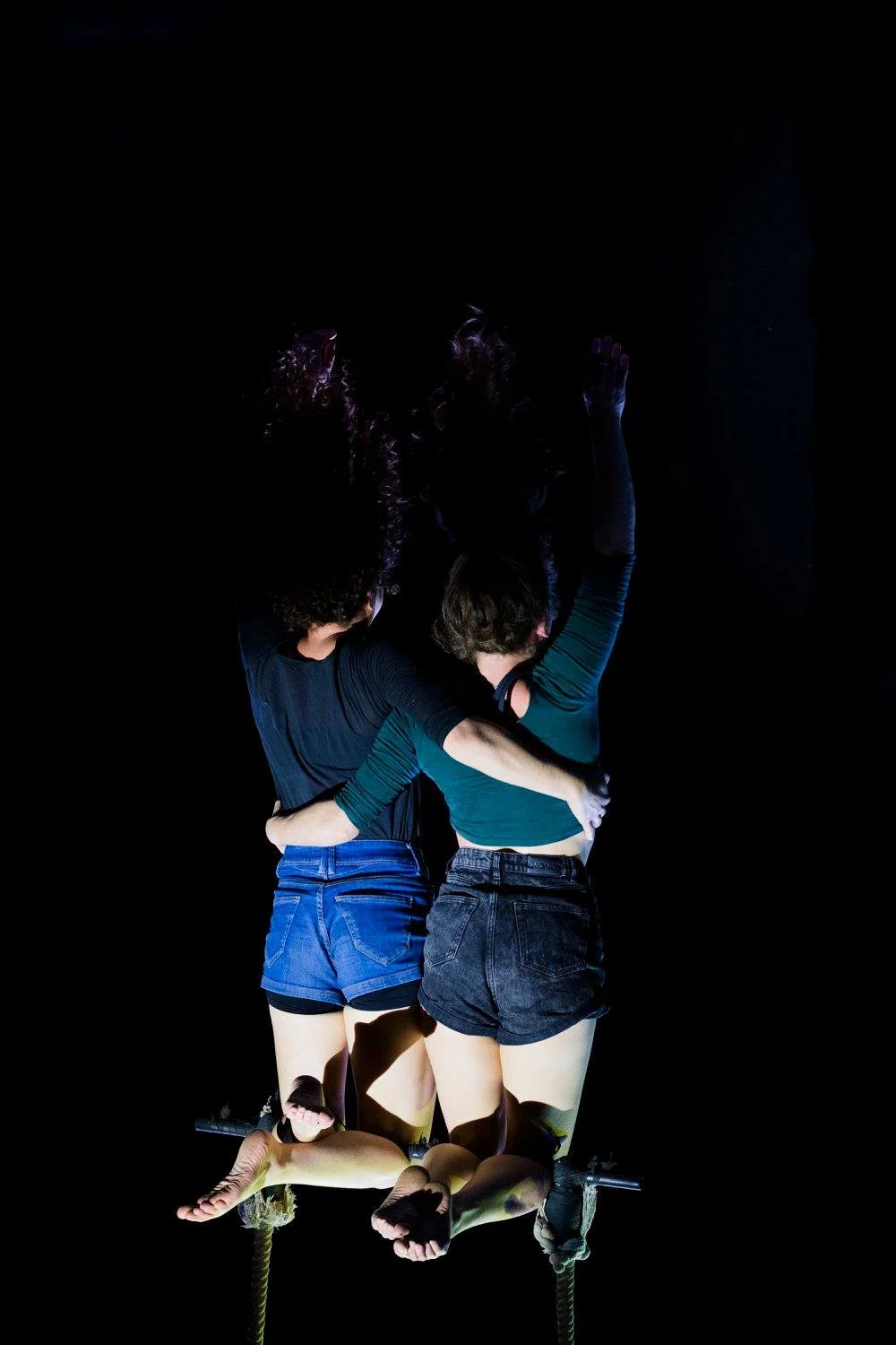 שתי אקרובטיות אוויר יושבות זו לצד זו על טרפז, זרועותיהן זו סביב זו והרגליים תלויות מול במה שחורה