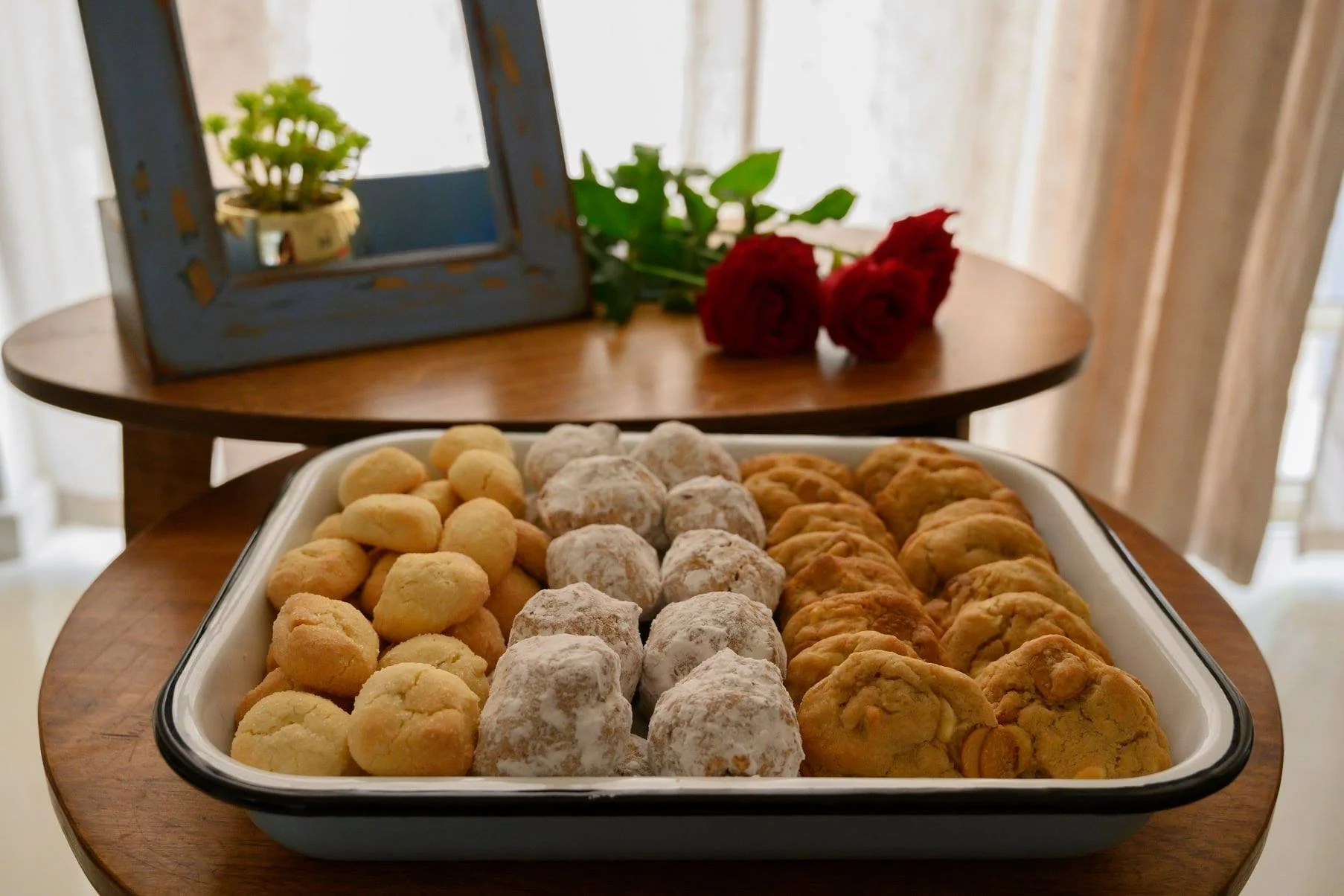 מגש עוגיות ביתיות ממוין לפי סוג על שולחן עץ, עם ורדים ותמונה ממוסגרת של צמח ברקע