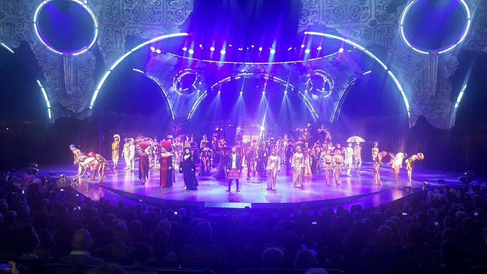 כל שחקני ההצגה קדים קידה על במת תיאטרון מוארת בכחול־סגול עם תפאורת קשת, קהל ישוב מלפנים