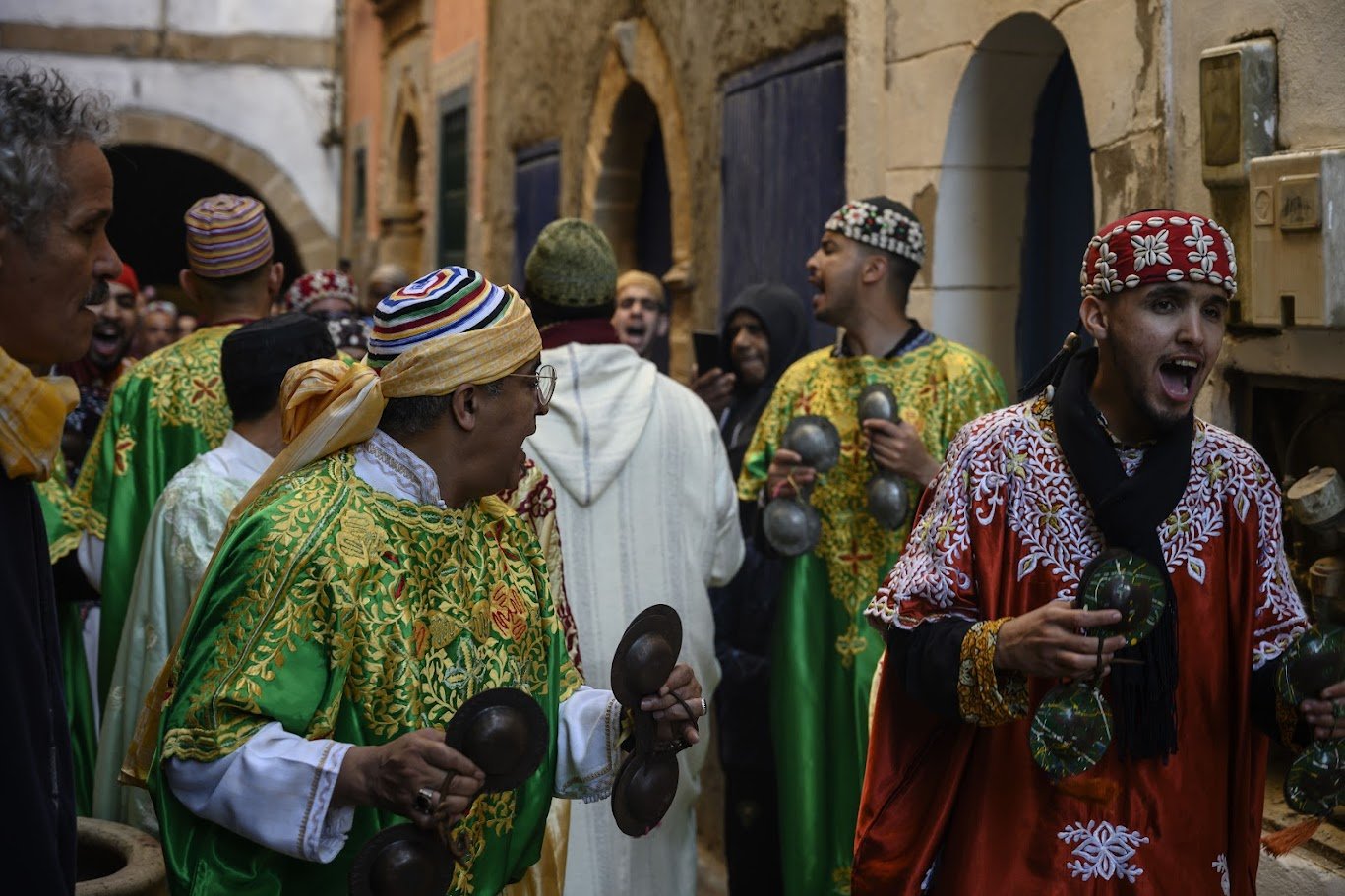 בסמטת עיר עתיקה צרה, מוזיקאים בגלימות רקומות מלווים חתונה מקומית בקסטנייטות מתכת ושירה – אסאווירה, מרוקו