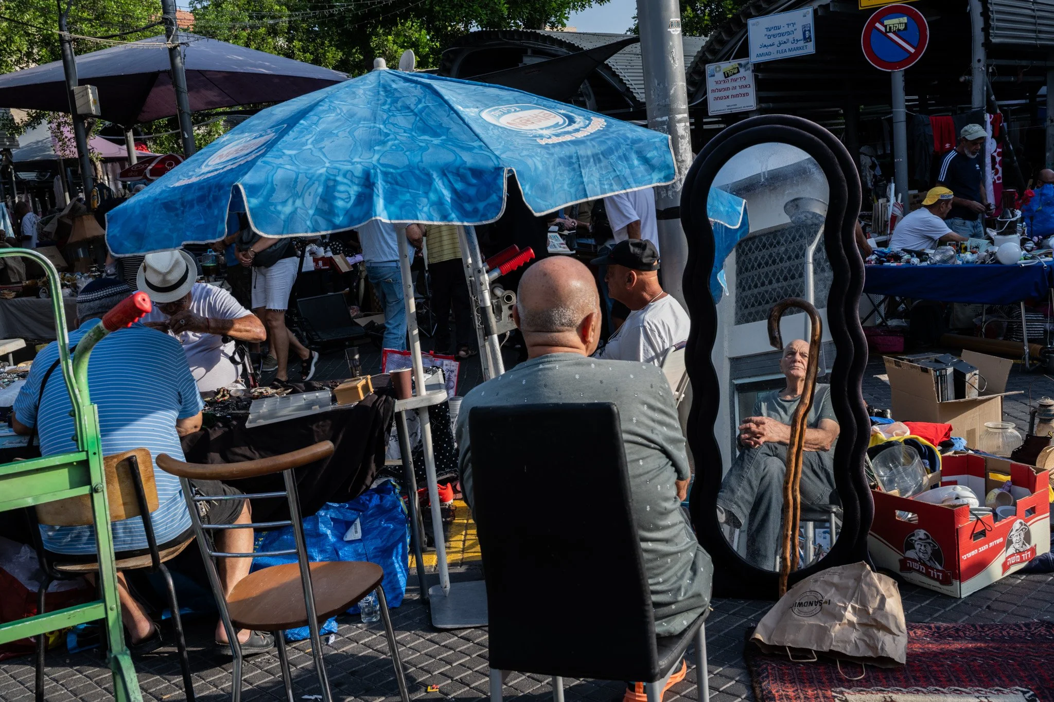 שוק פשפשים פתוח מתחת למטריות כחולות עם אנשים יושבים ומראה גלית גבוהה שמשקפת את הסצנה