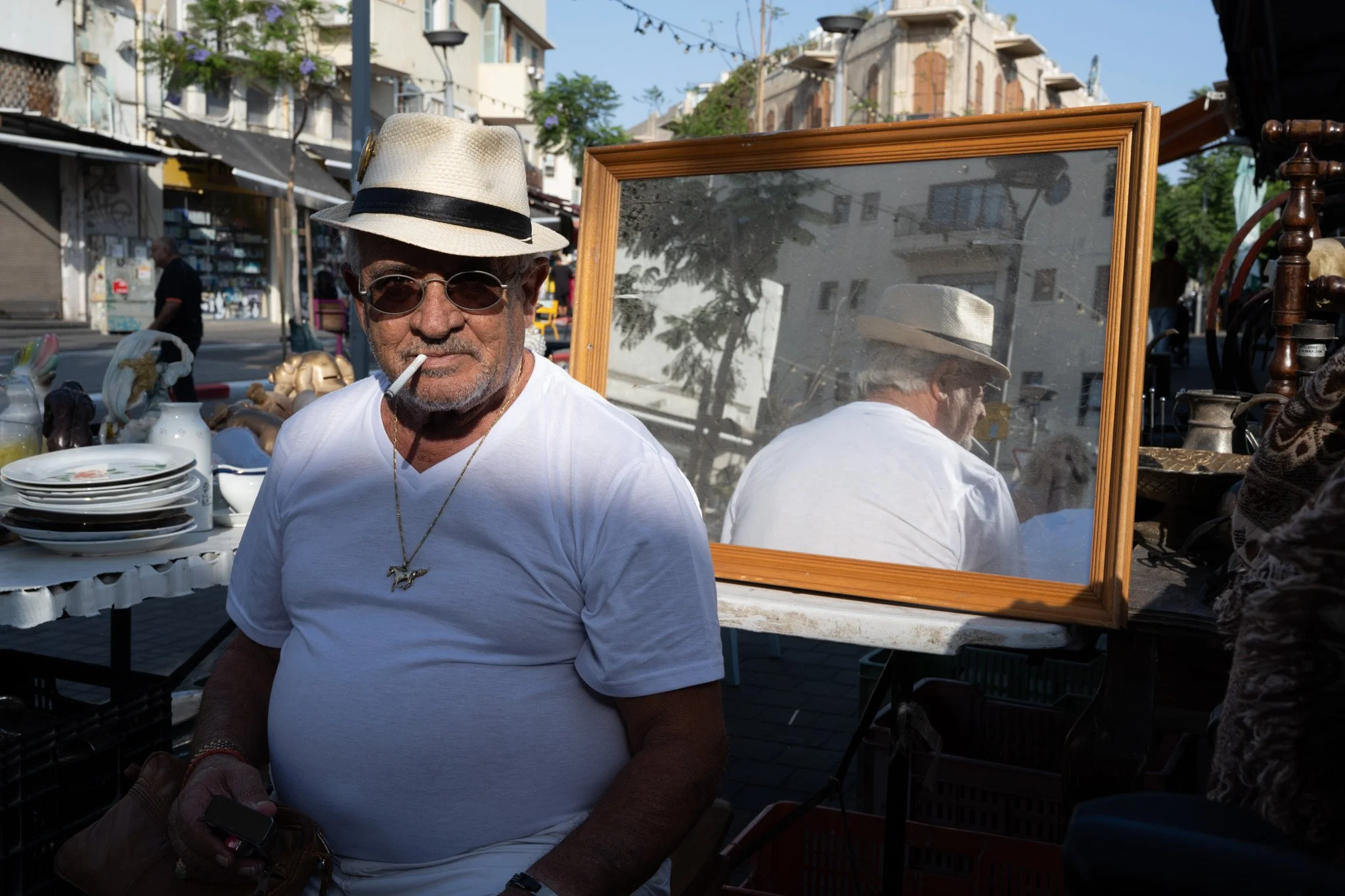 סוחר רחוב בחולצה לבנה וכובע קש עומד ליד מראת תצוגה בשוק הומה – יפו, ישראל