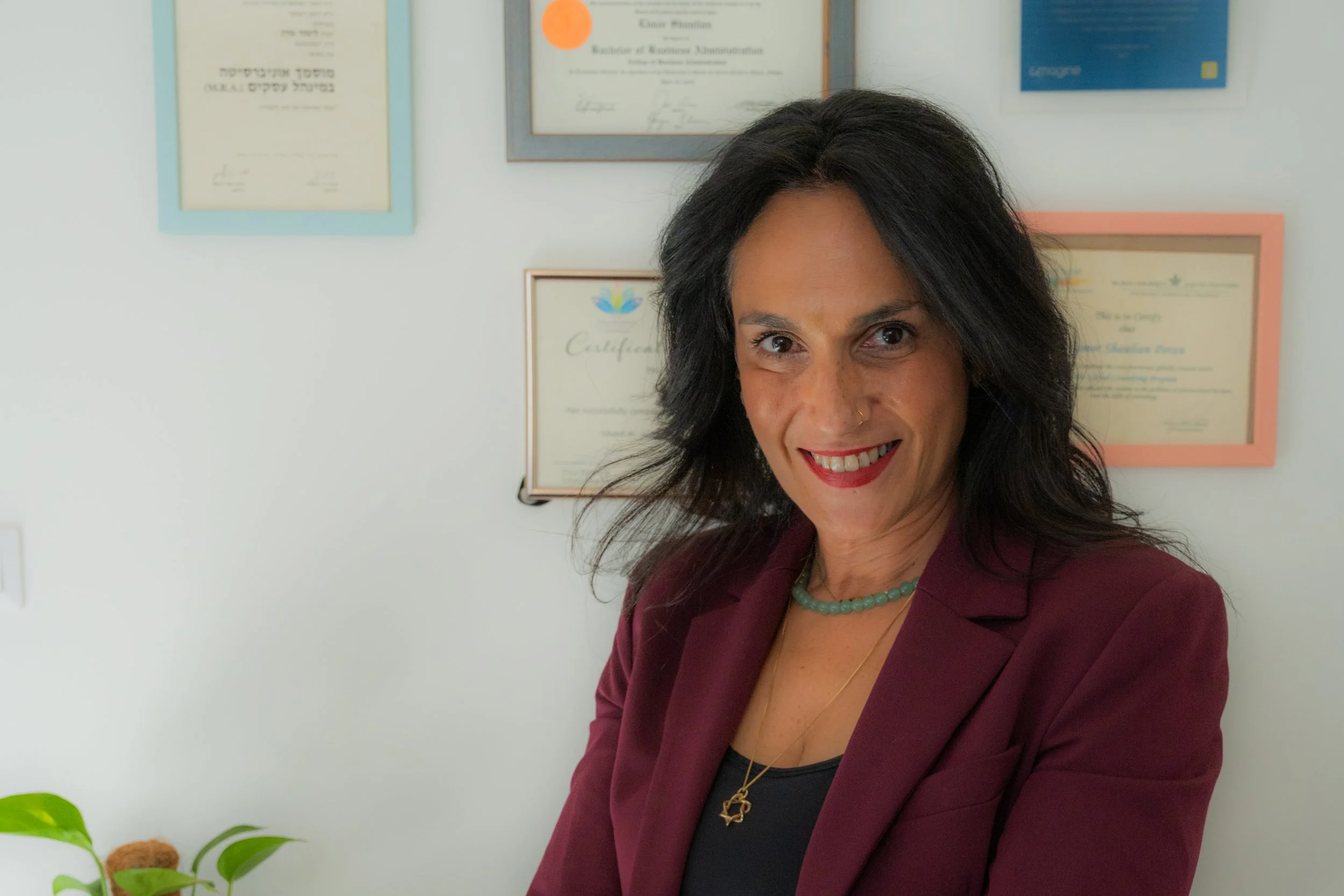 Limor Poran sitting confidently in front of a wall of diplomas and certificates, wearing a burgundy blazer and jeans, blending warmth and professionalism.