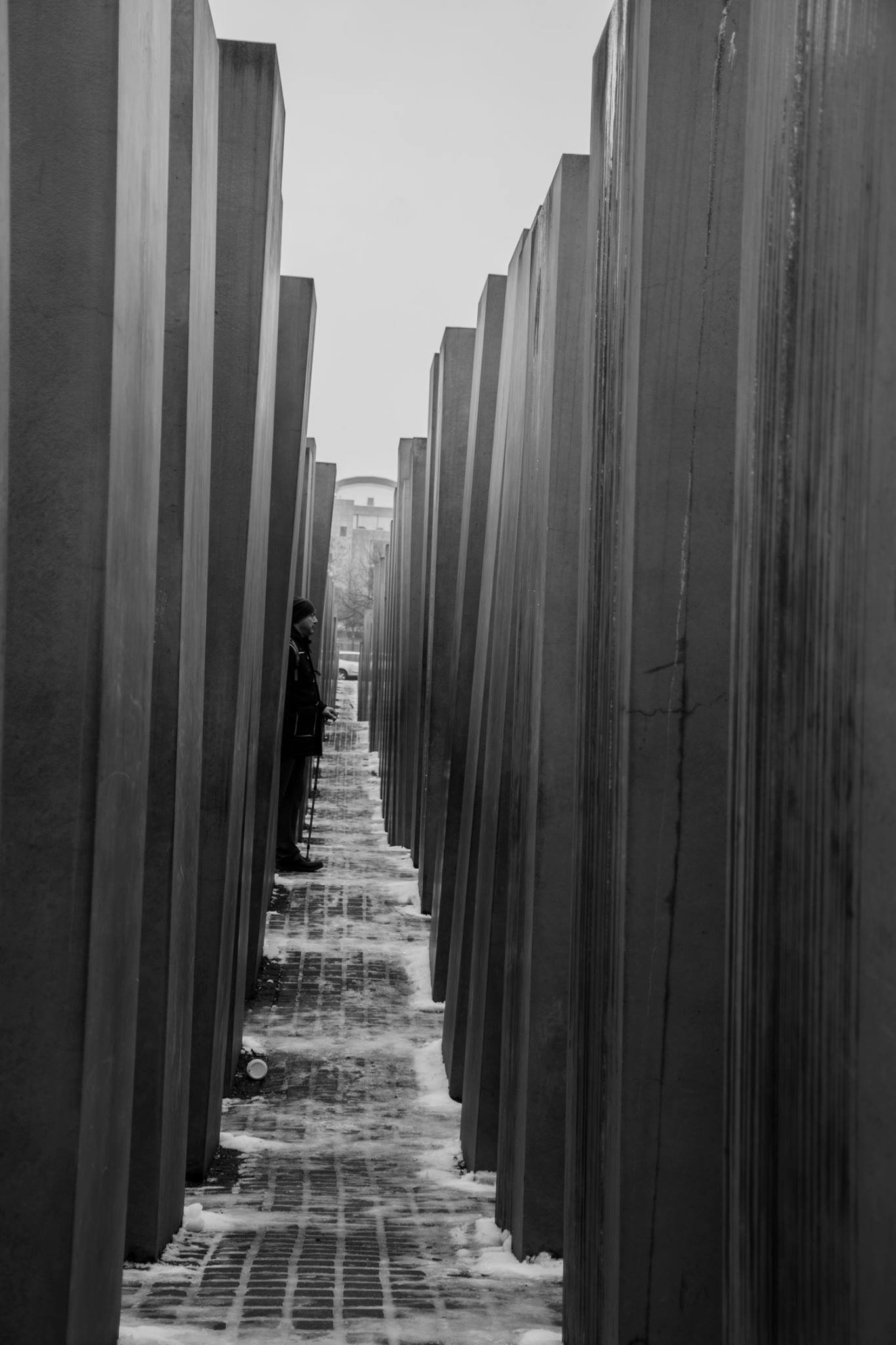 מבט בשחור־לבן לאורך המסדרונות הצרים של אנדרטת השואה בברלין, מבקר יחיד ושלג תחת הרגליים