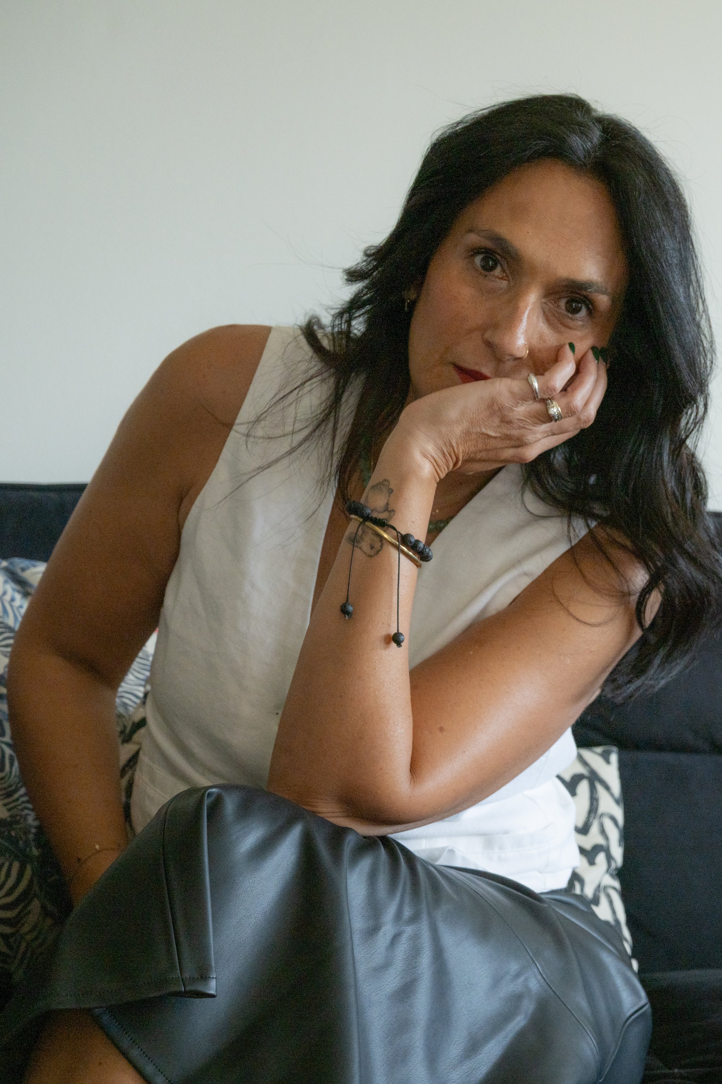 Limor Poran sitting on a dark sofa, head resting on her hand, looking directly at the camera with a thoughtful, vulnerable expression