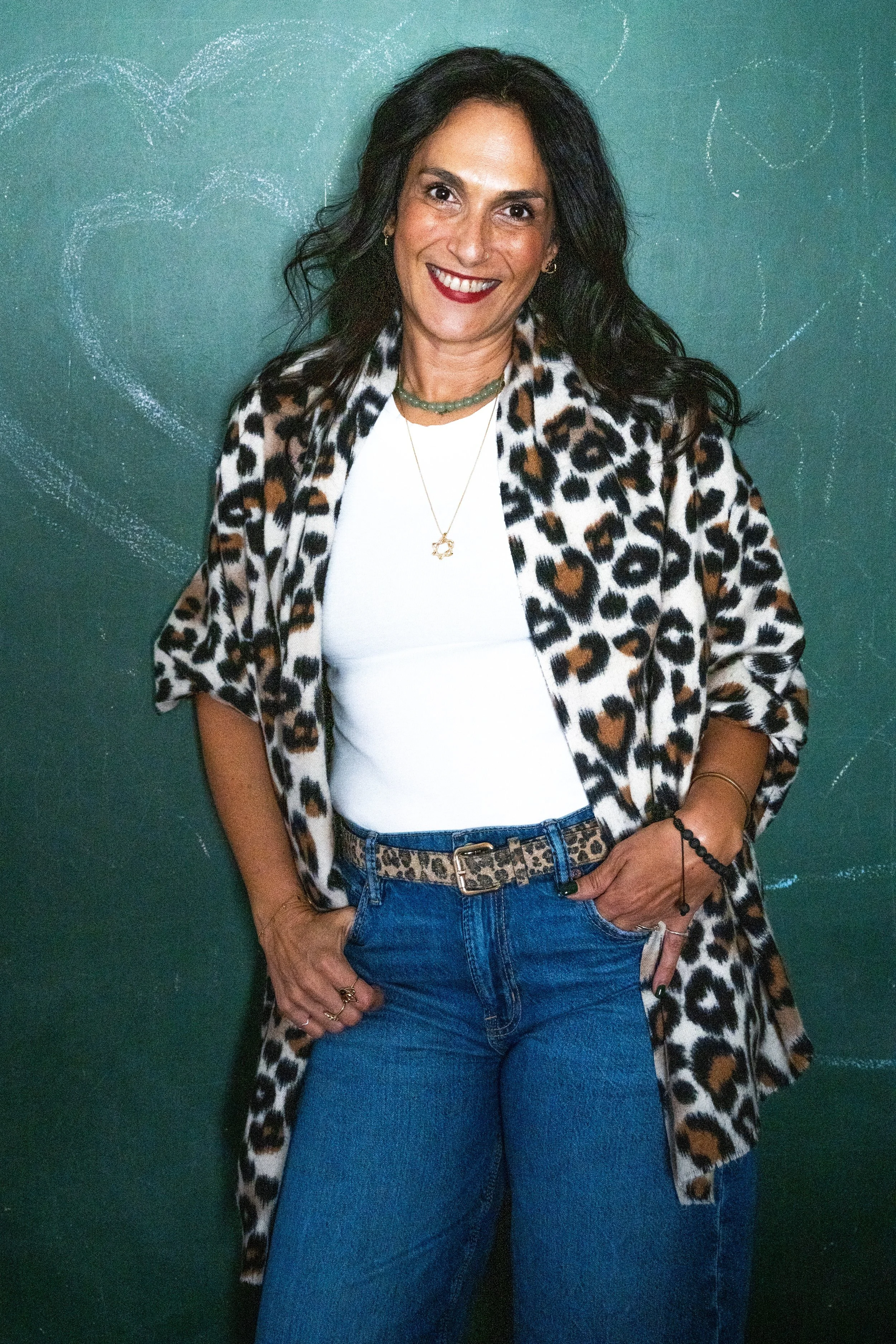 Limor Poran in a leopard-print cardigan and jeans, smiling in front of a chalkboard with faint heart drawings, radiating playful confidence