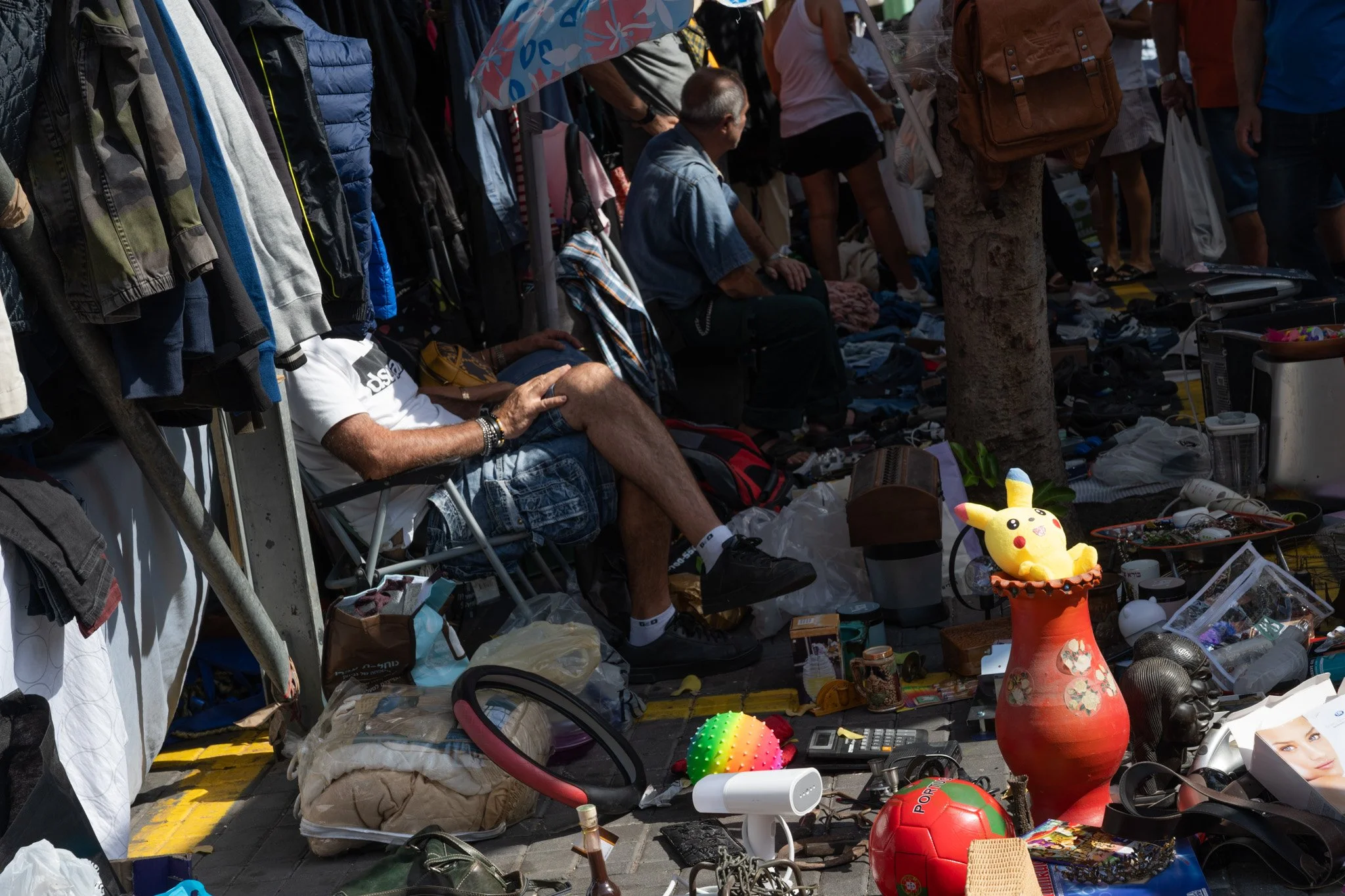 דוכן בשוק הפשפשים בגובה הרצפה עם רגליו של המוכר היושב בין חפצים עמוסים, צעצועים, הגאים ואגרטל אדום